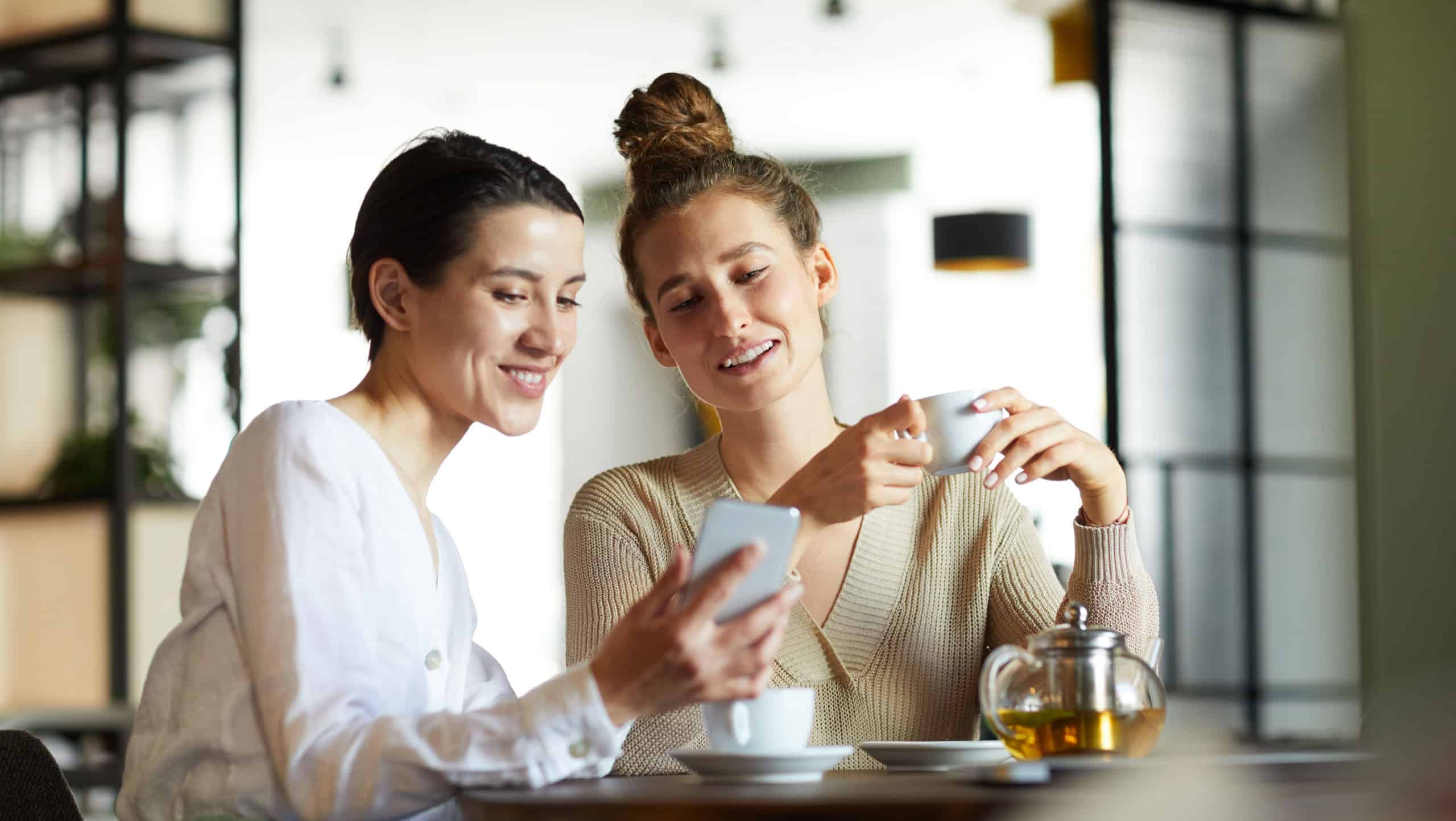 Two young females in casualwear watching curious video in smartphone while having tea in cozy cafe