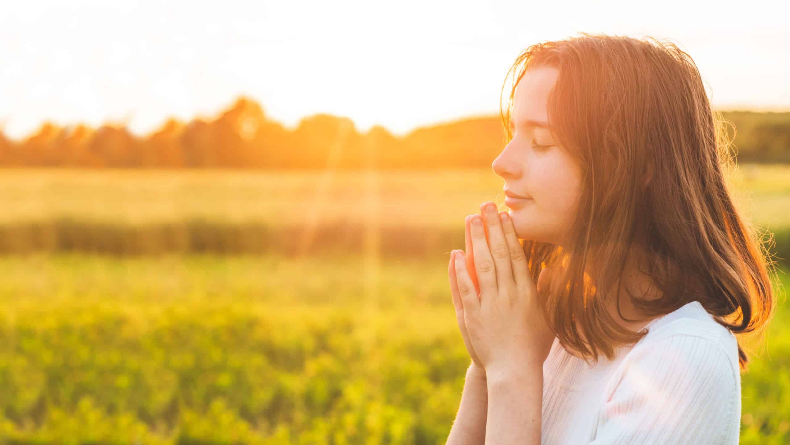 Teenager Girl closed her eyes, praying in a field during beautiful sunset. Hands folded in prayer concept for faith, spirituality and religion. Peace, hope, dreams concept