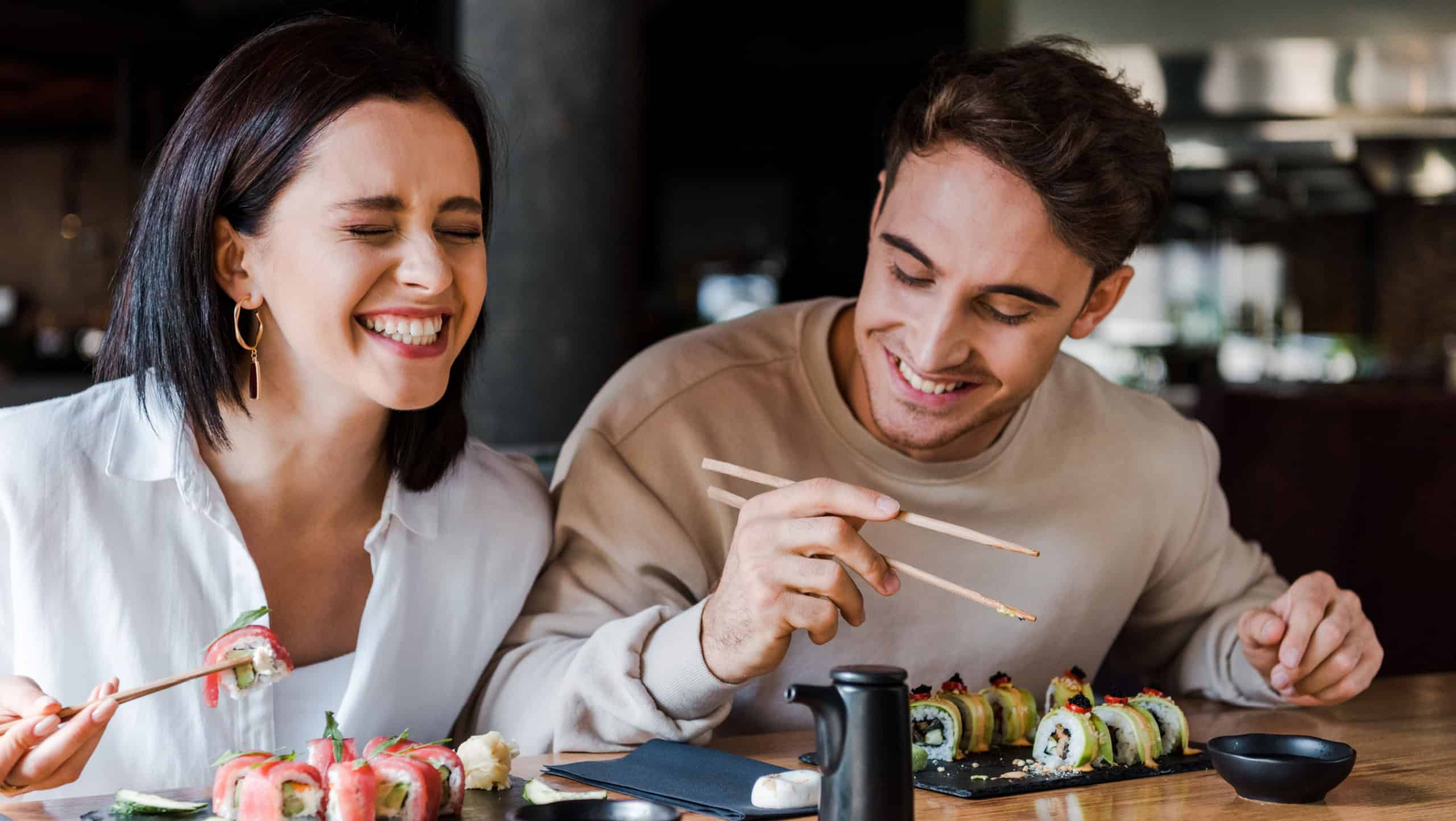 happy man and cheerful woman laughing while holding chopsticks with tasty sushi in restaurant