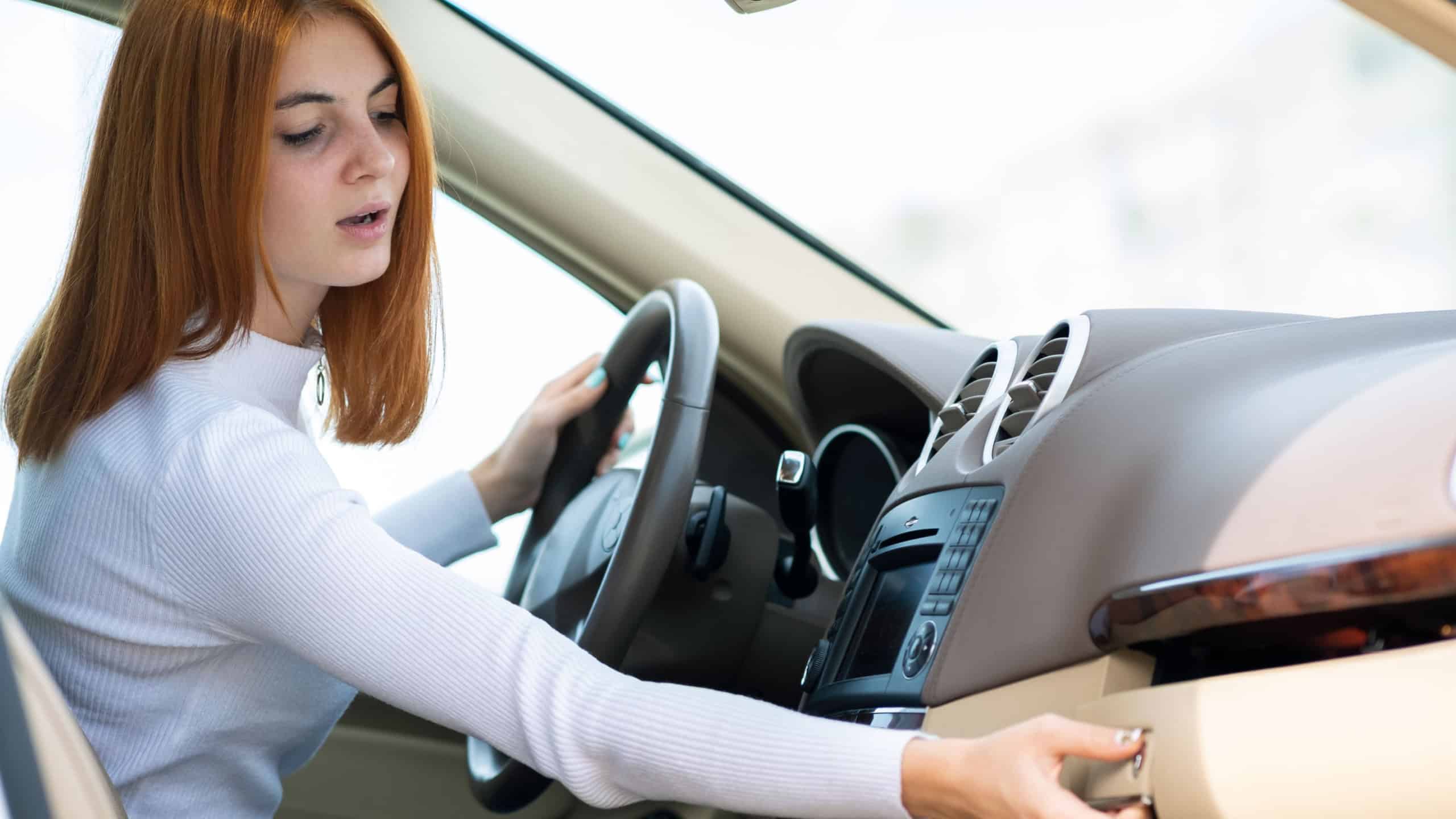 Young red haired woman driving a car and disctucted while looking for something in glove compartment.