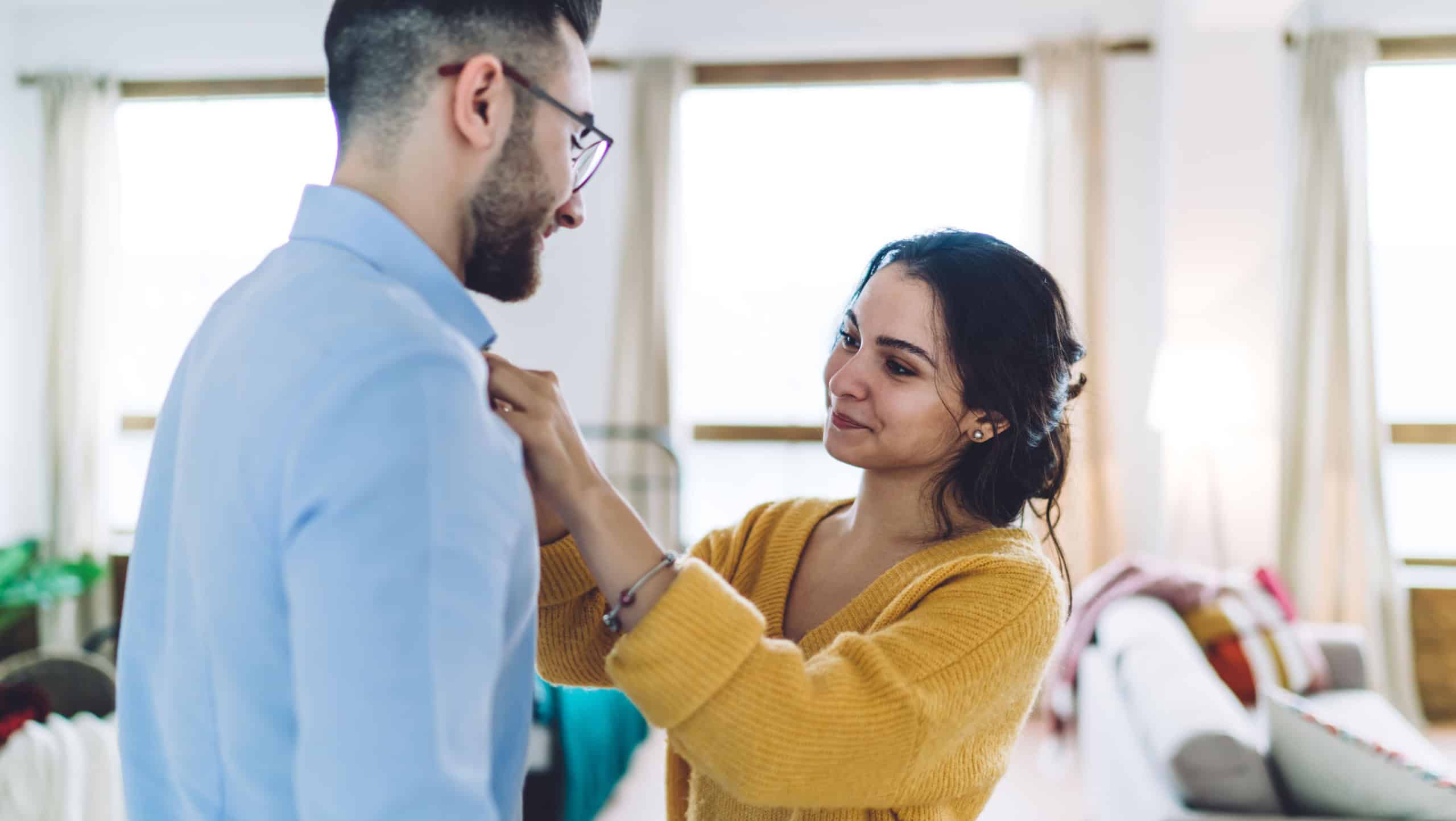 Side view of smiling young female in casual yellow jumper taking care of beloved husband and fixing collar before leaving for work