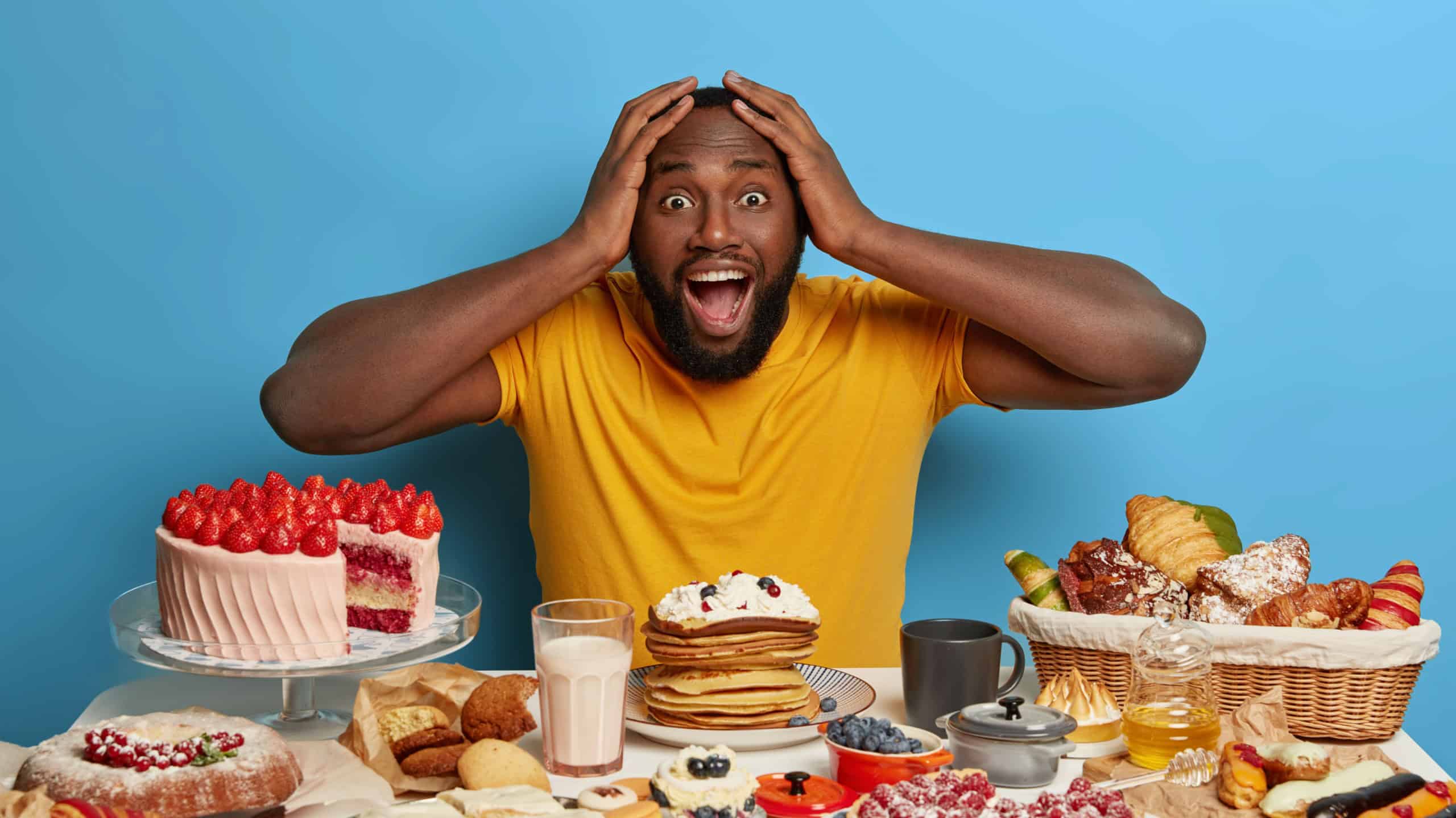 Emotive dark skinned bearded guy keeps both hands on head, opens mouth from surprisement as sees too much desserts at one table, shocked his wife baked lots of confectionery for festive event
