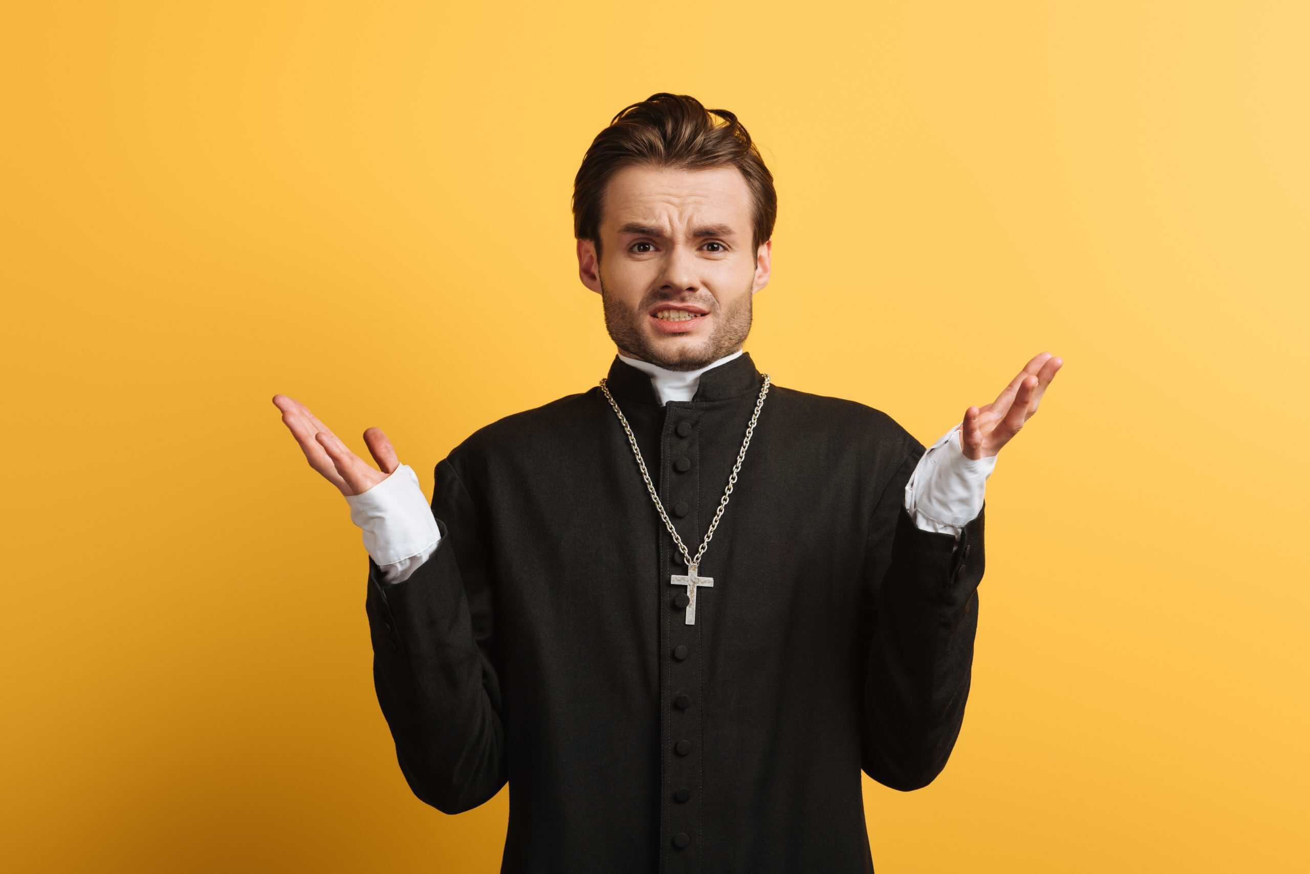 Discouraged catholic priest showing shrug gesture at camera isolated on yellow