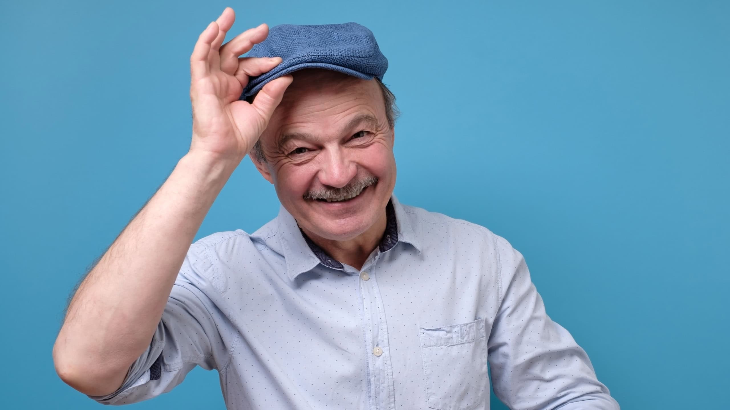 Cheerful senior man greets by taking off a hat. Studio shot on blue wall.