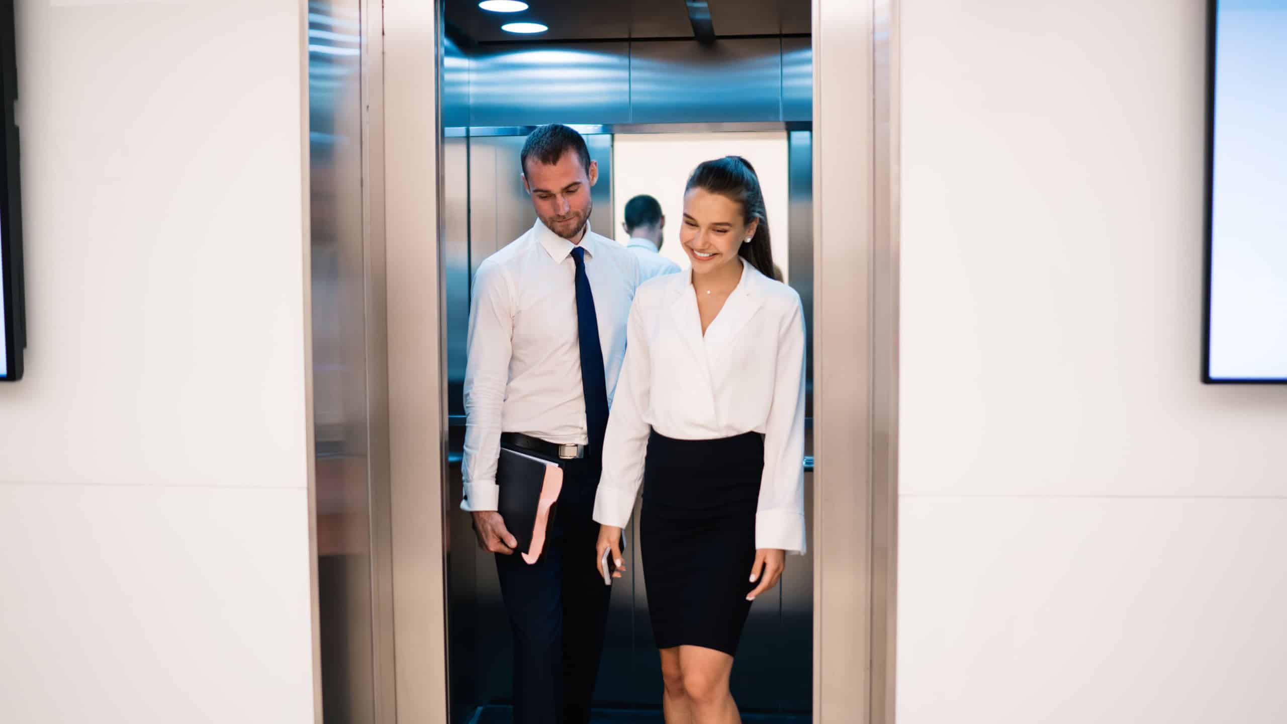 Young couple of male carrying black folder and female carrying smartphone in official wear smiling and walking out of lift together into contemporary bright workspace