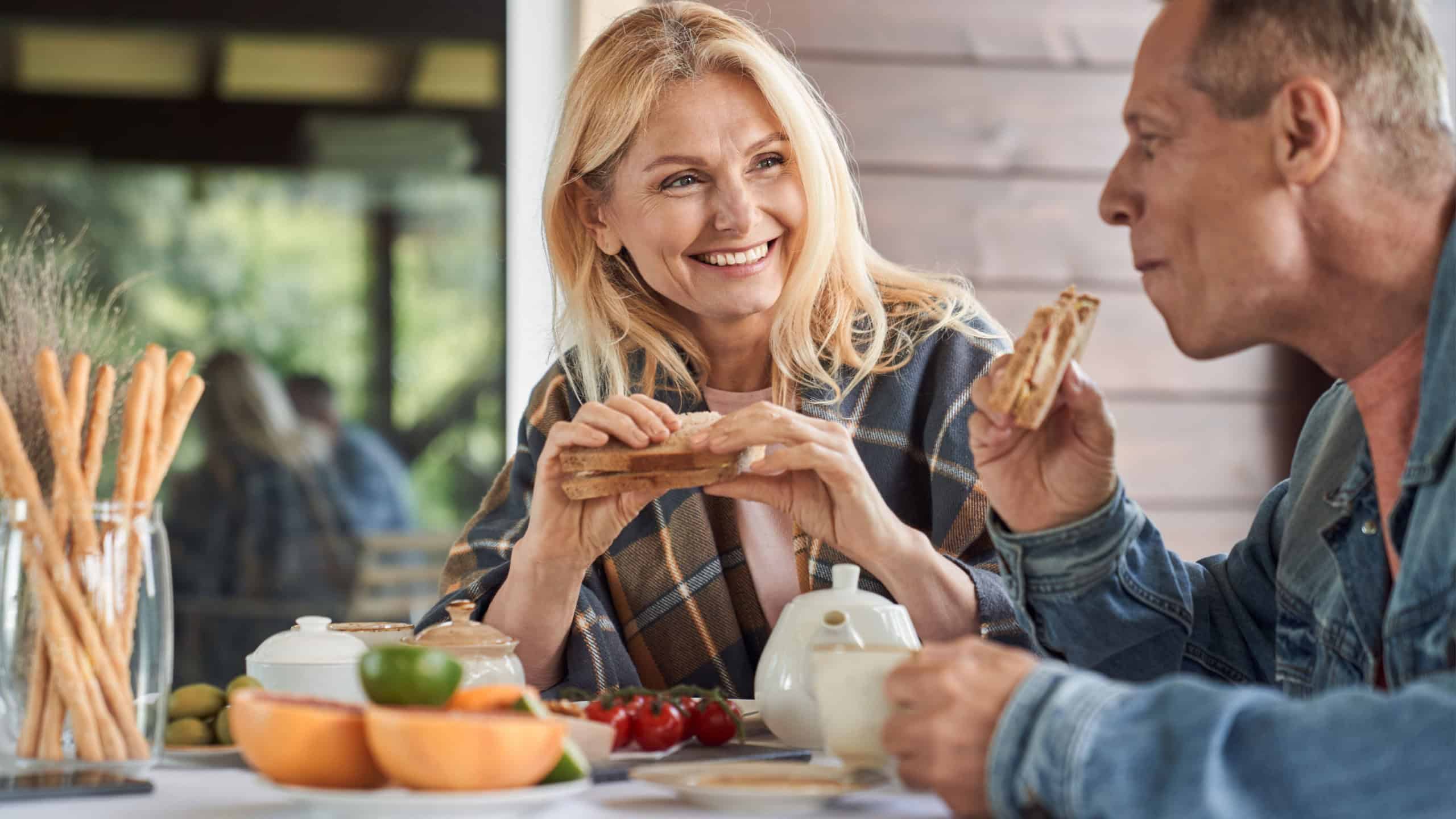 Smiling mature man and woman are having toasts with vegetables and hot drinks on terrace in countryside
