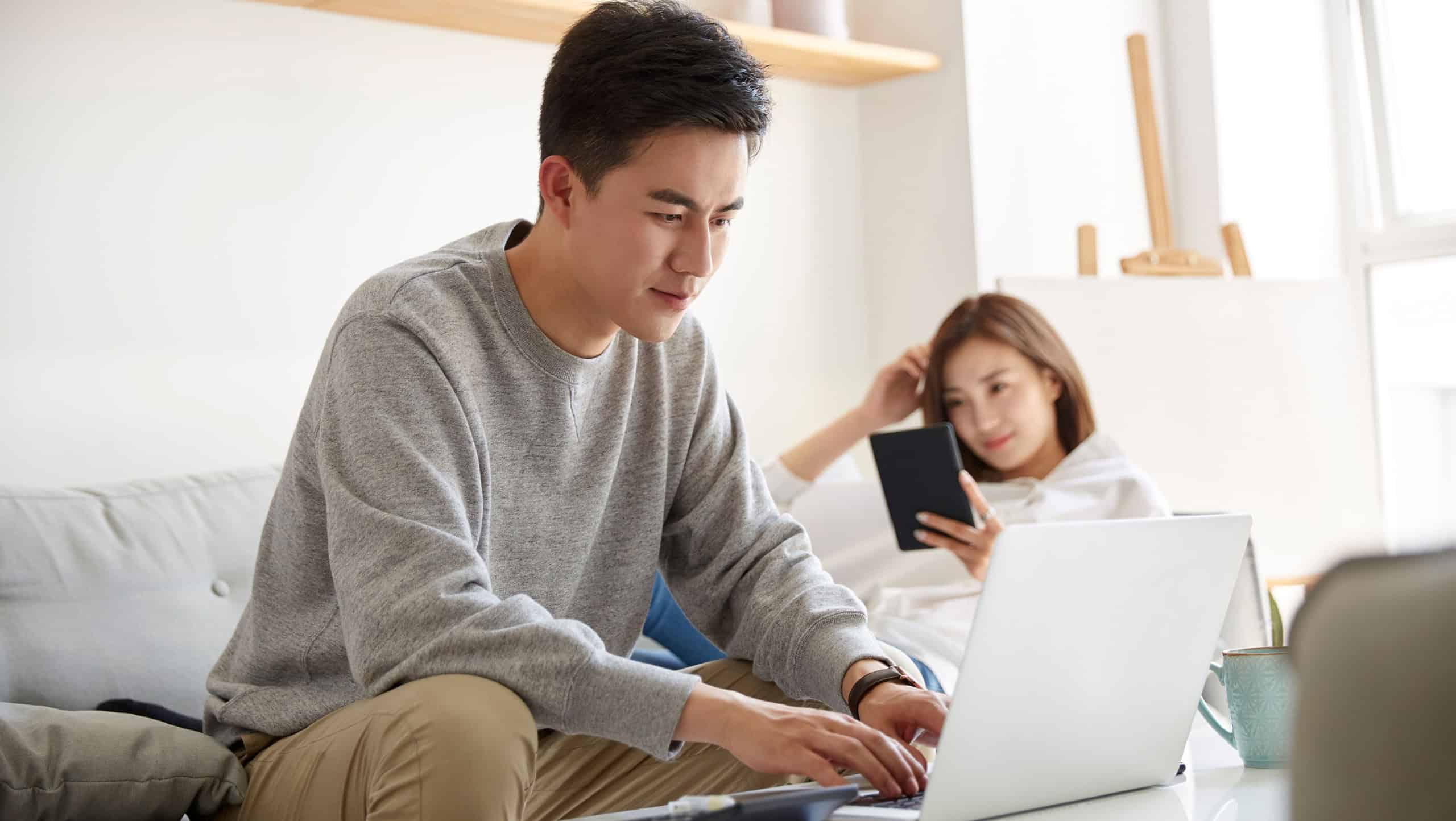 young asian couple staying at home with husband working using laptop computer and wife reading ebook