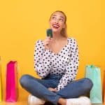 What To Buy. Portrait of trendy woman holding credit card and looking up at copy space, thinking about purchase, dreaming about new clothes, sitting on the floor at yellow studio background
