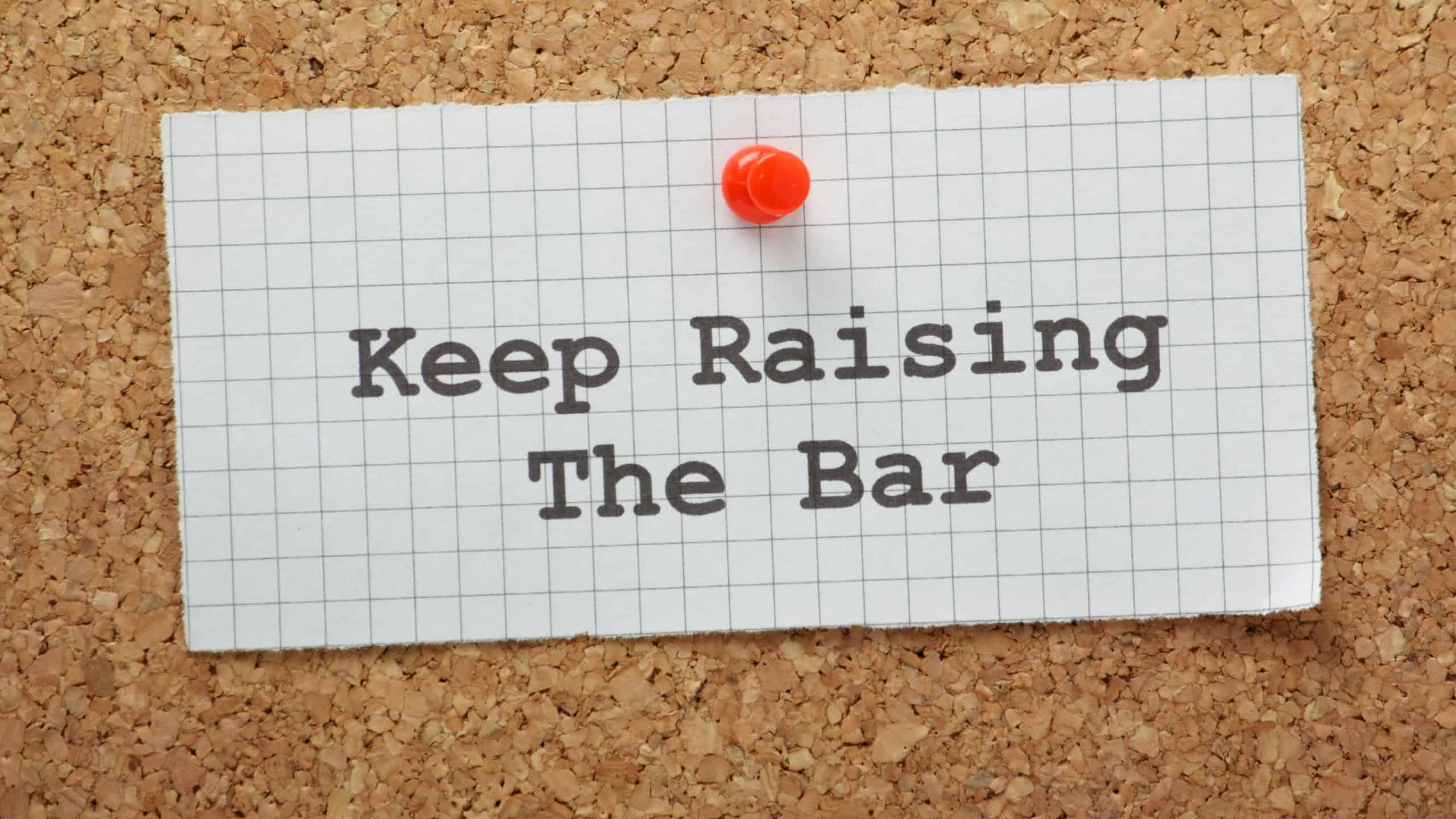 The phrase Keep Raising the Bar on a piece of graph paper pinned to a cork notice board. A concept for raising standards and setting targets as a form of motivation.