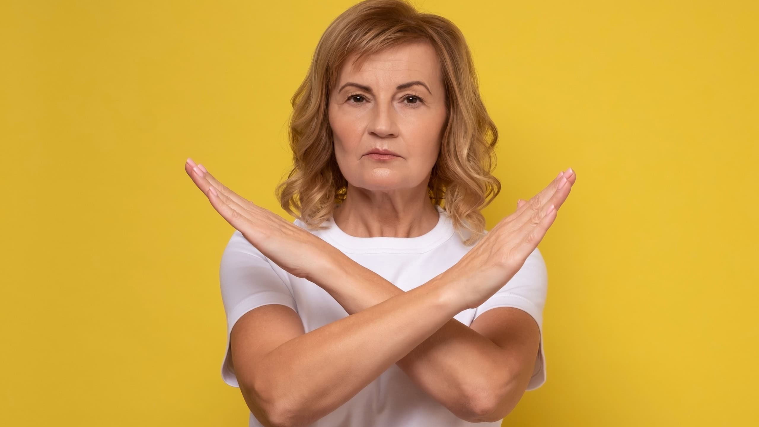 Mature woman making X sign with crossed hands, gesturing stop, warning of danger. No way, stop doing. Studio shot on yellow background