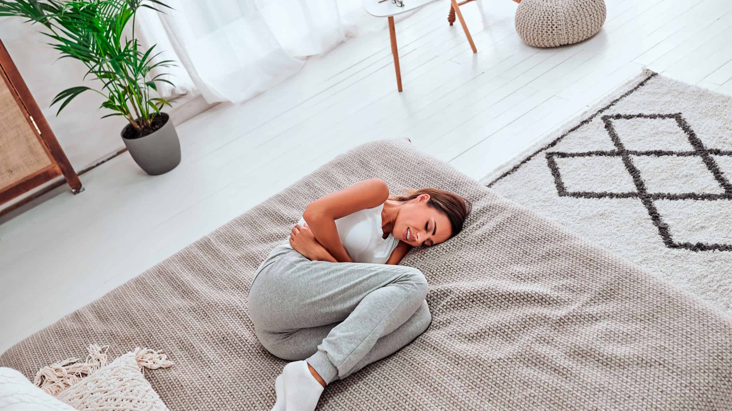 Young beautiful woman in painful expression holding her belly suffering menstrual period pain lying sad on home couch having tummy cramp in female health concept. Top view.