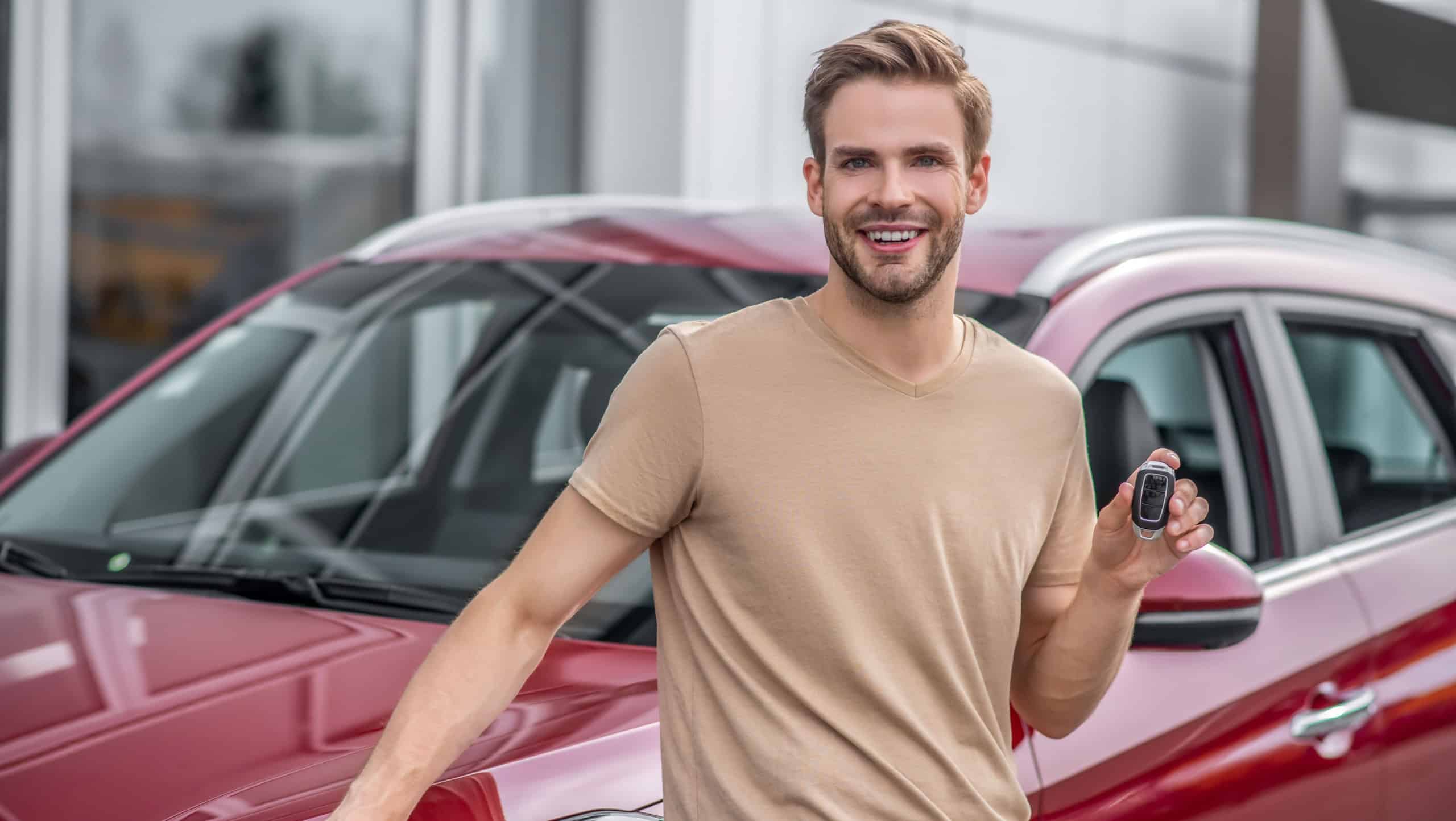 Brand-new car. Smiling young male leaning on red car, holding keys