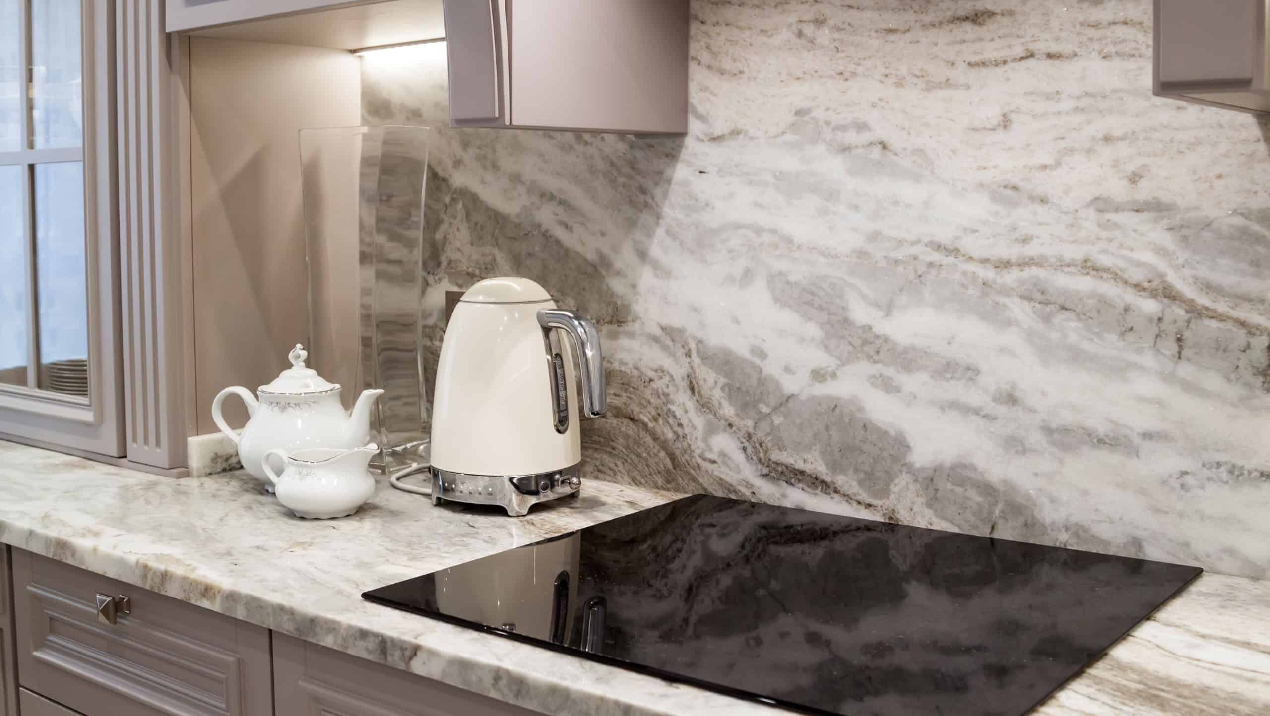 beautiful closeup of custom designed kitchen, with marble looking quartz countertop and backsplash. cream electric kettle with porcelain tea accessories on the marble countertop next to ceramic hob