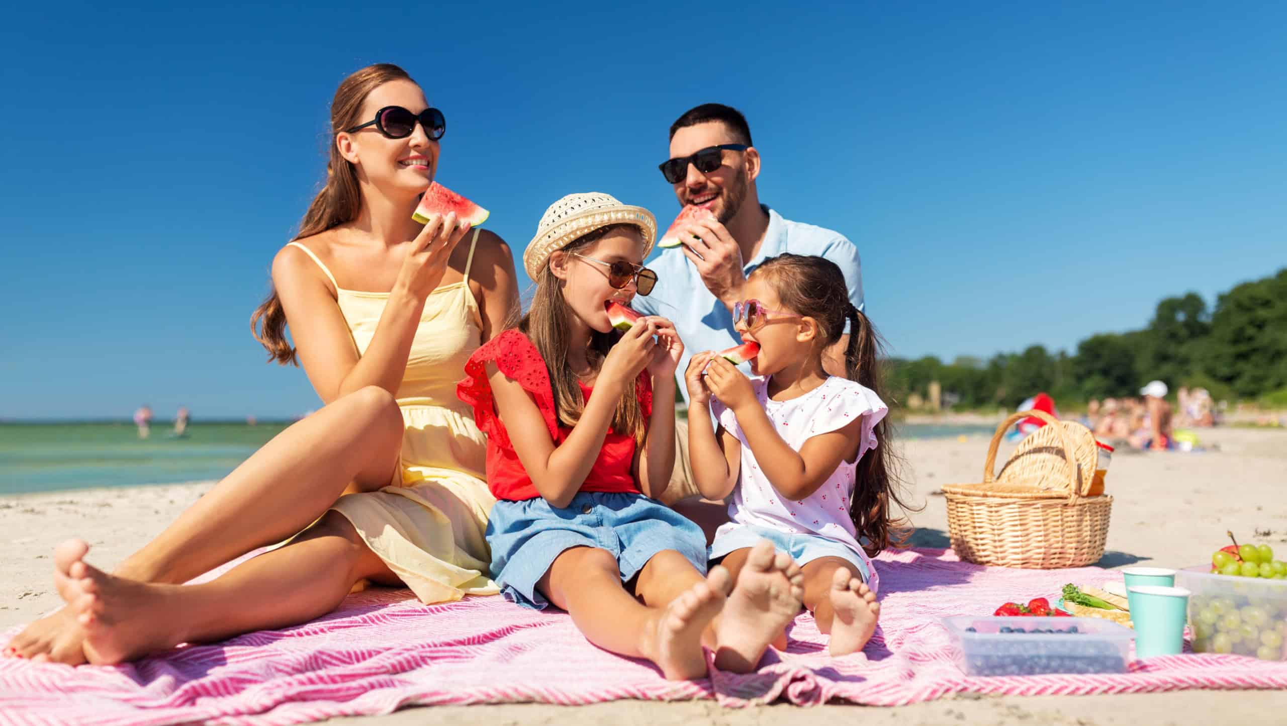 family, leisure and people concept - happy mother, father and two daughters having picnic on summer beach and eating watermelon