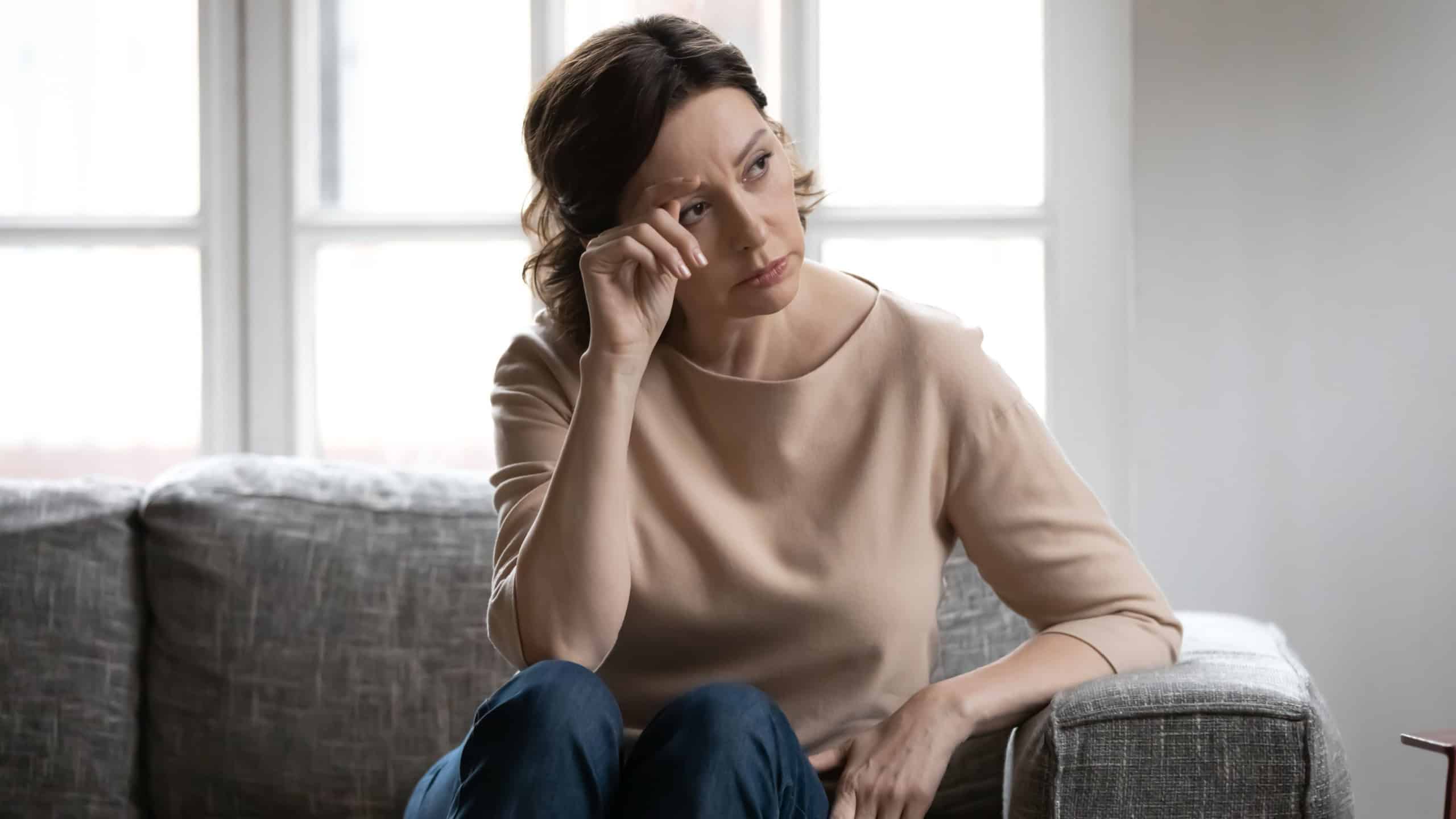 Thoughtful mature woman touching forehead, lost in thoughts, memories, sitting on couch at home alone, upset pensive middle aged female looking in distance, thinking about problems, pondering