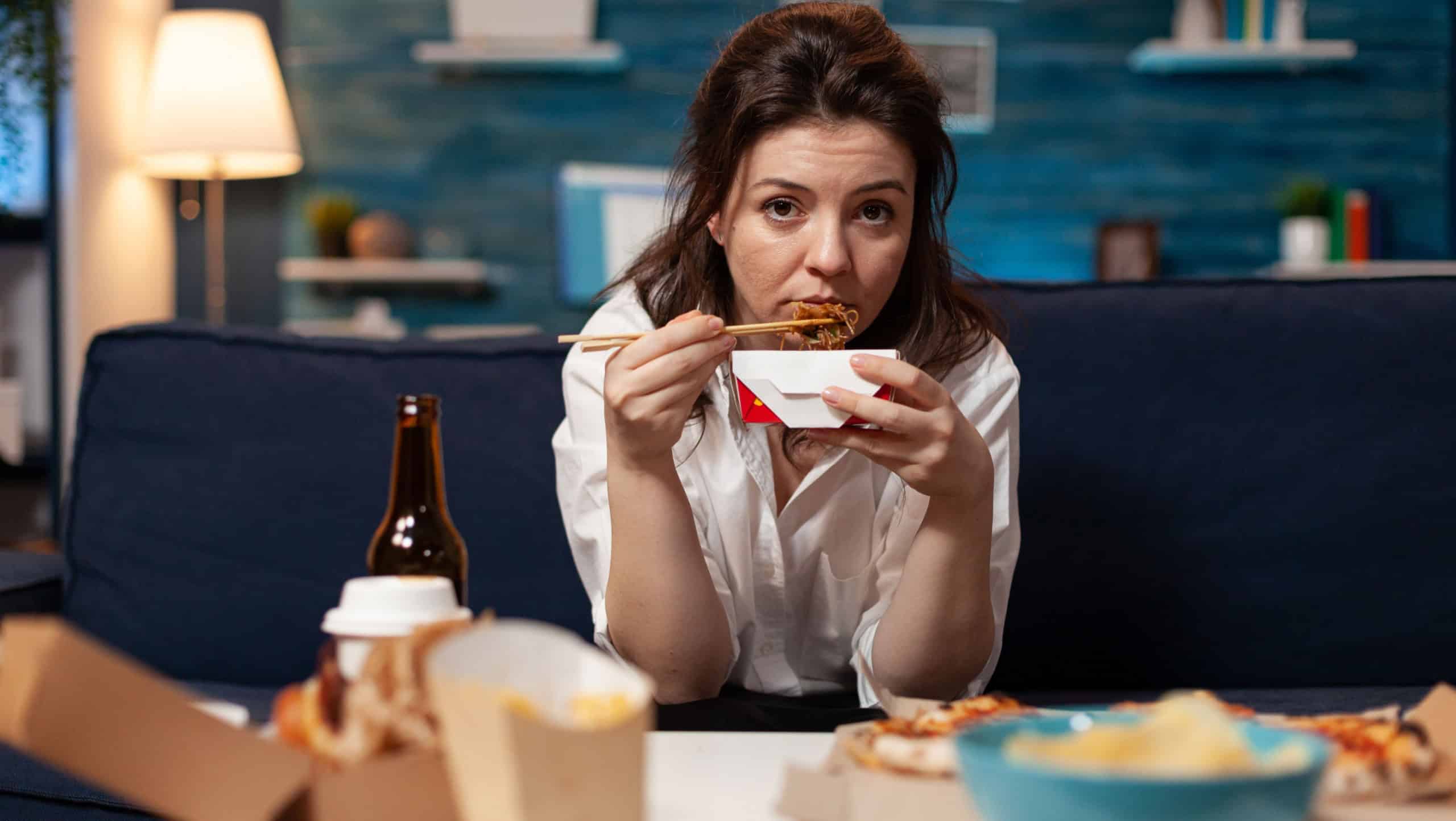 Portrait of caucasian female looking into camera while eating chinese chopstick relaxing on sofa in living room late at night. Woman enjoying takeaway food home delivered. Tasty fastfood lunch order