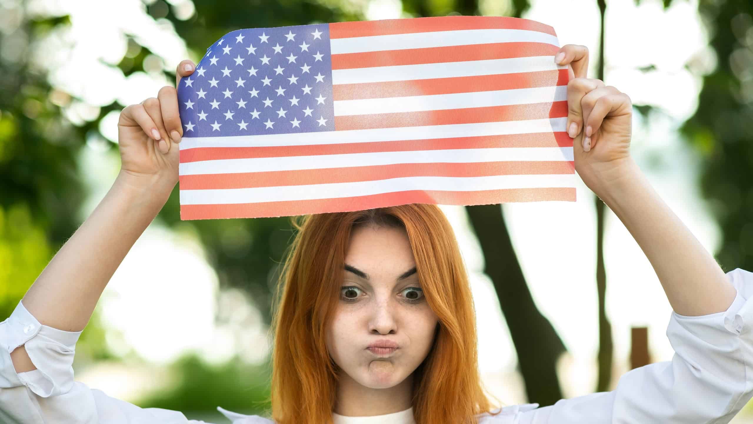 Angry young red haired woman protester posing with USA national flag up over her head standing outdoors in summer park. Activist girl on demonstration with national banner.