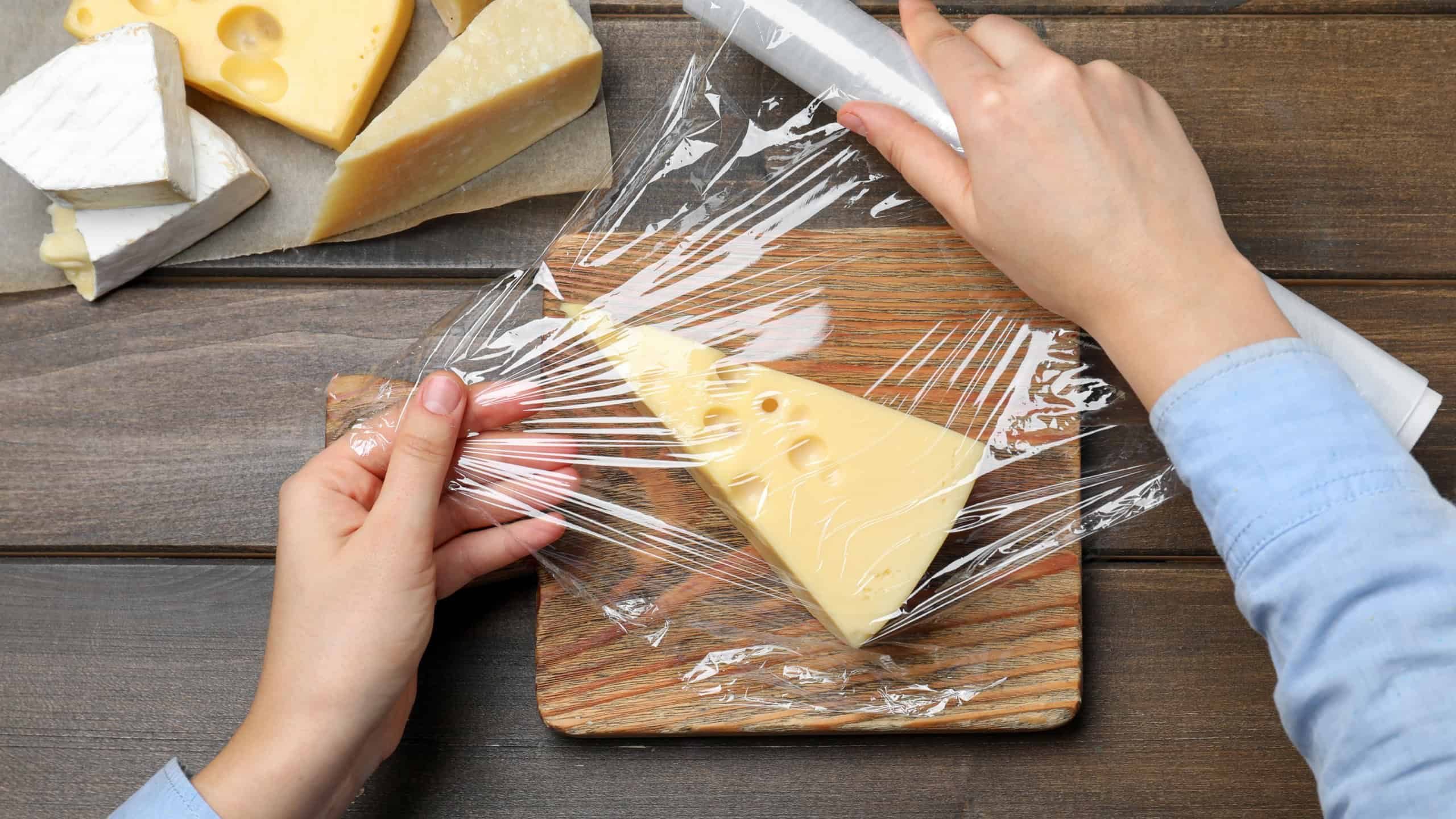 Woman putting plastic food wrap over block of cheese at wooden table, top view