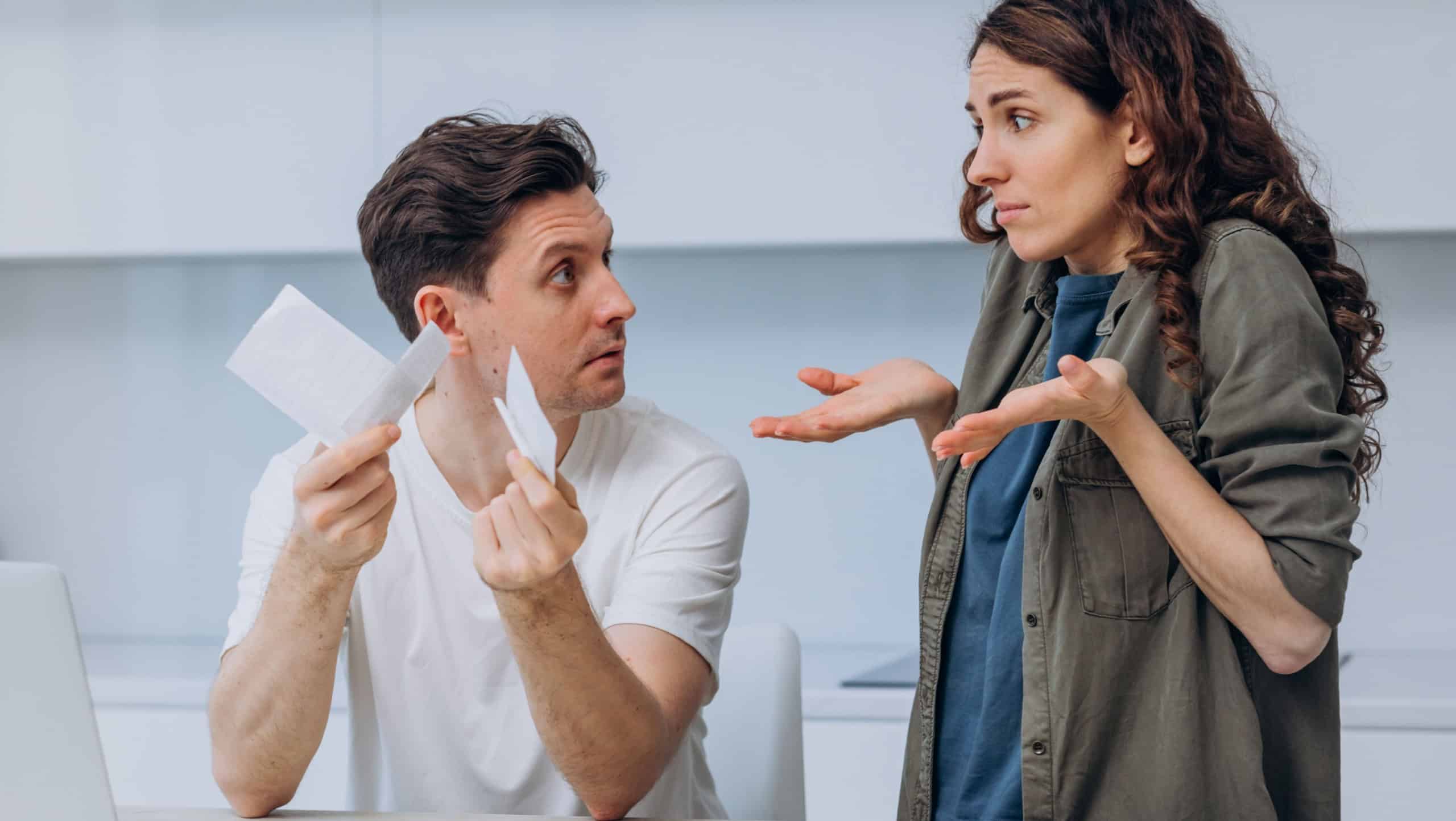 Husband shows his wife expensive checks and asks where the money went while sitting at a laptop in the kitchen, the wife throws up her hands in confusion and does not know what to answer