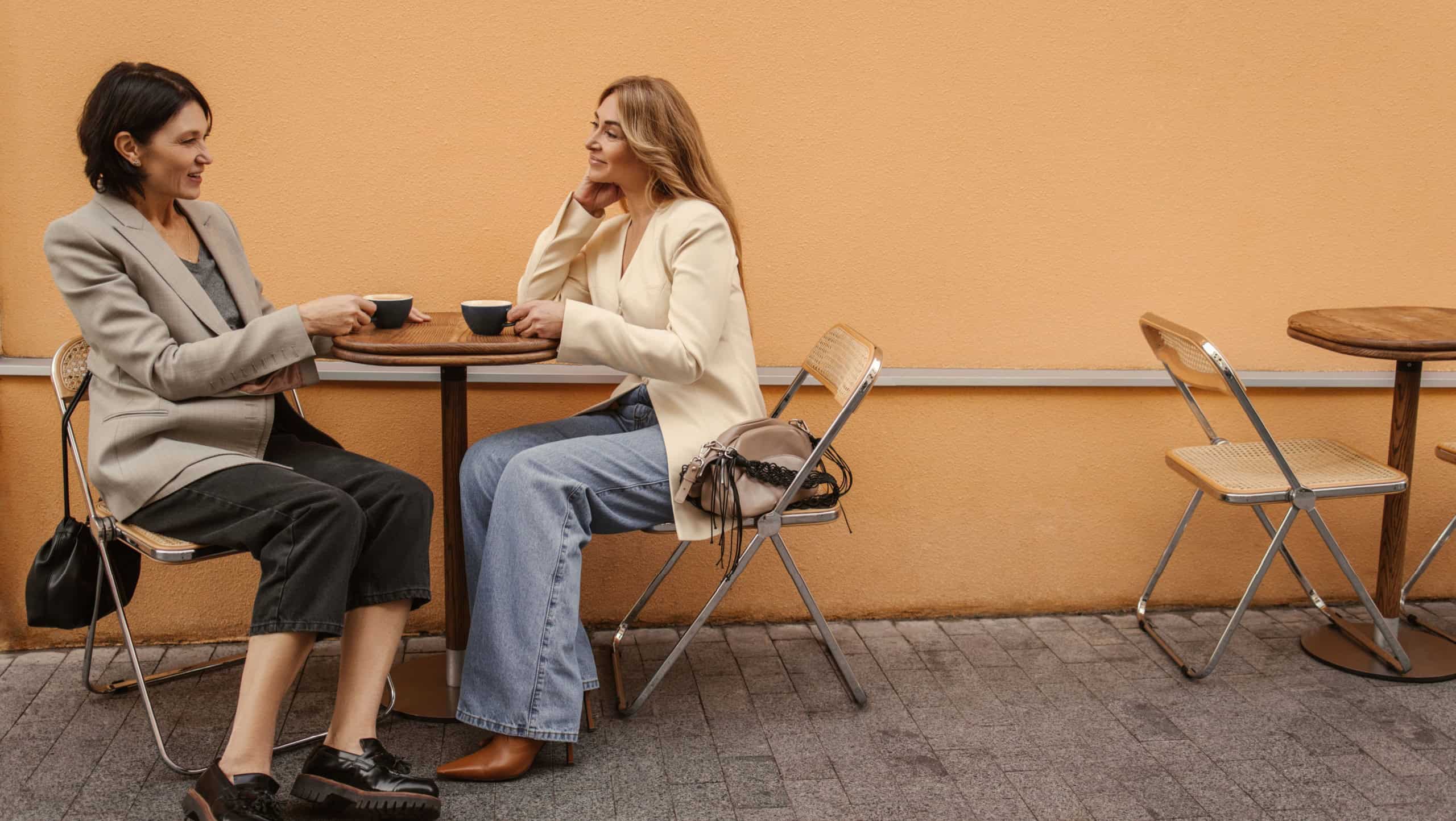 Two middle-aged caucasian ladies are sitting at table on street, chatting and drinking coffee, tea. Brunette and blonde in casual clothes are having relaxed time. Lifestyle and people concept
