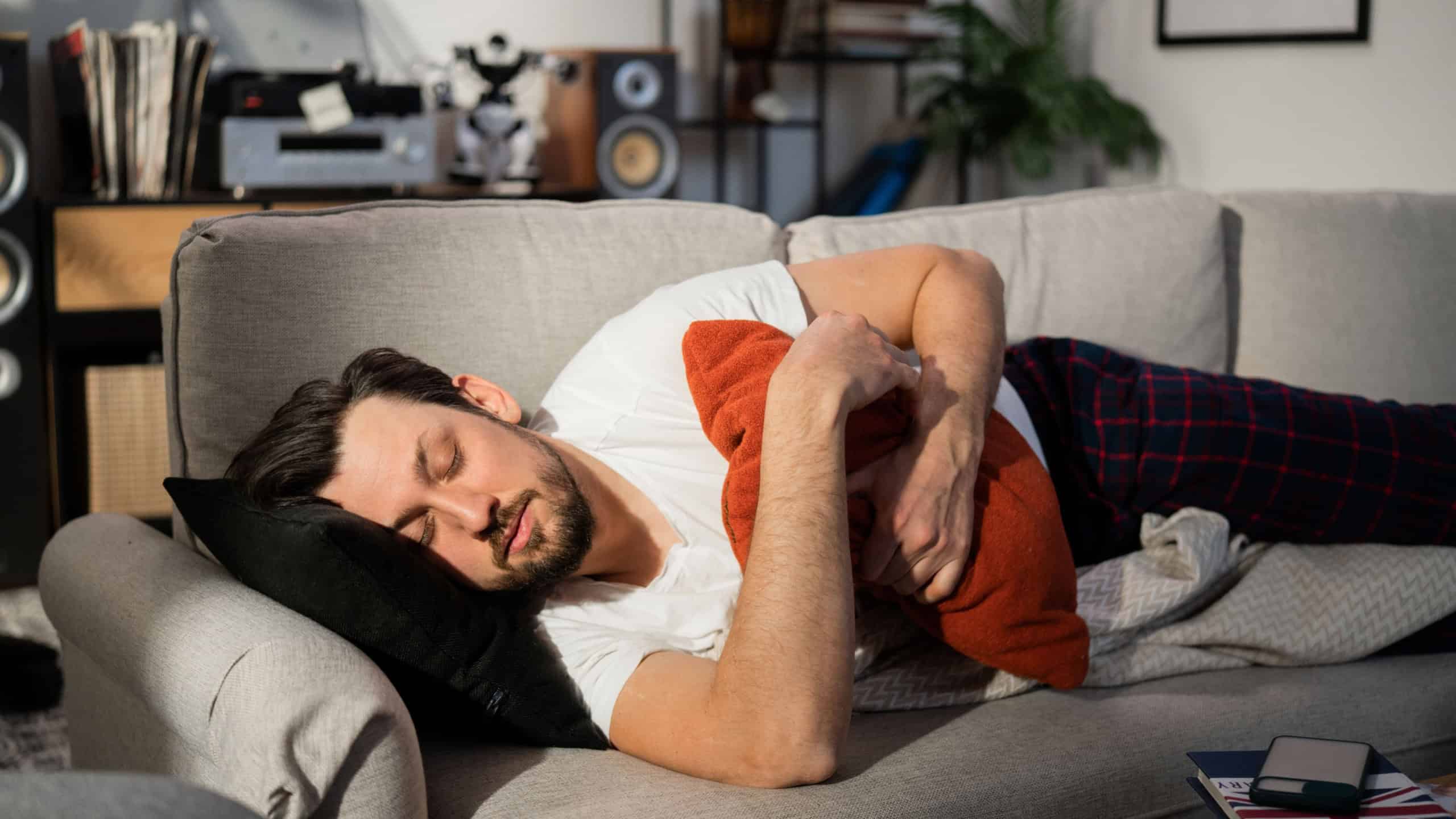 Young with beard falls asleep on the couch in the living room. The sun shines on his face. The man hugged pillow with hands. The brunette is dressed in pajamas and lying on blanket.
