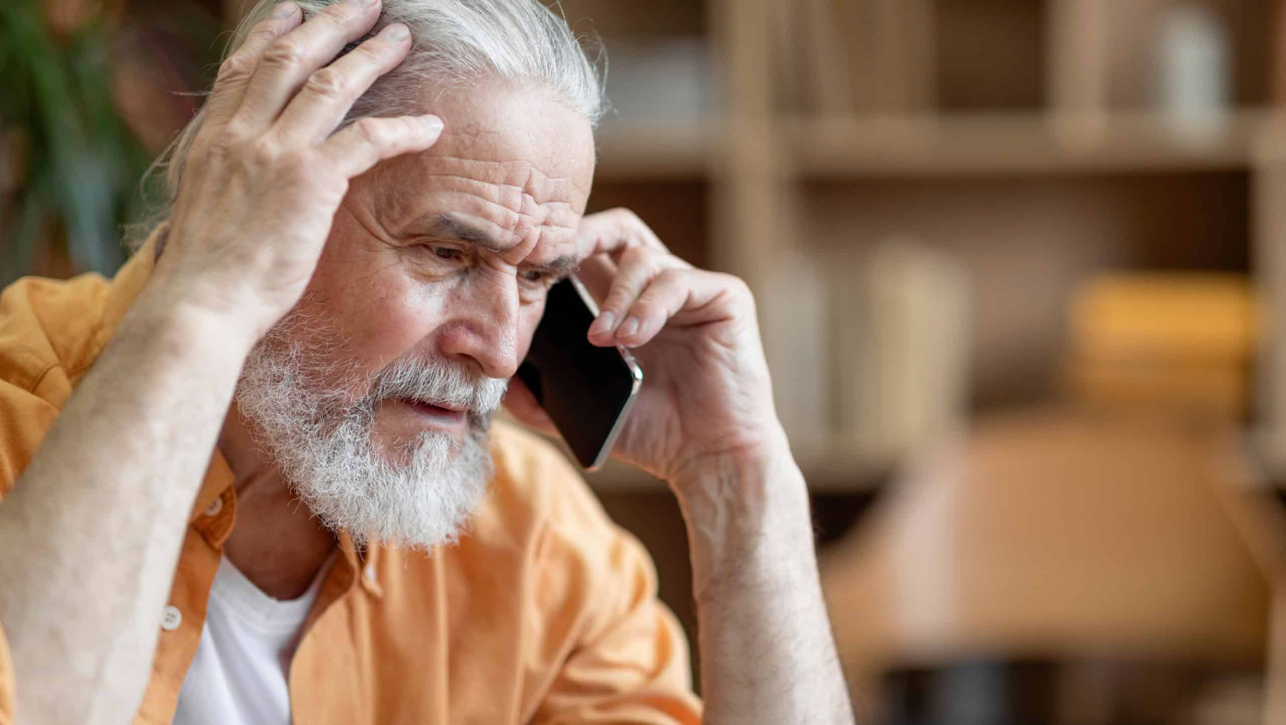 Unhappy senior man having phone conversation with someone, touching his head and looking down, upset grandfather getting bad news, home interior, copy space, closeup shot