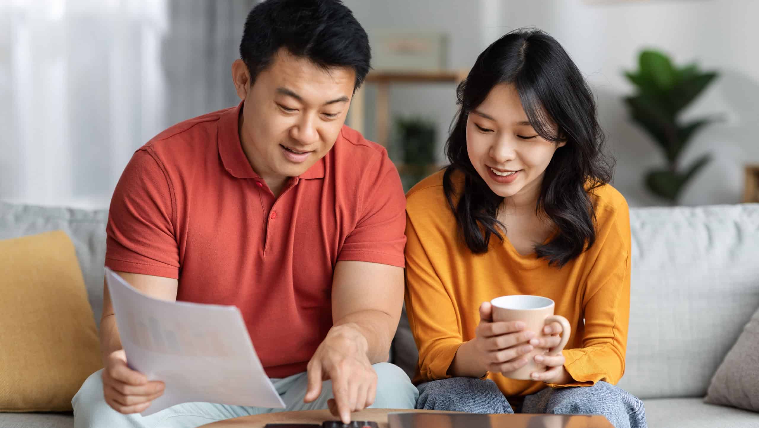 Happy asian family middle aged husband and young wife sitting on couch, holding documents, calculating monthly spending, tracking expenses at home, satisfied with result, family budget concept