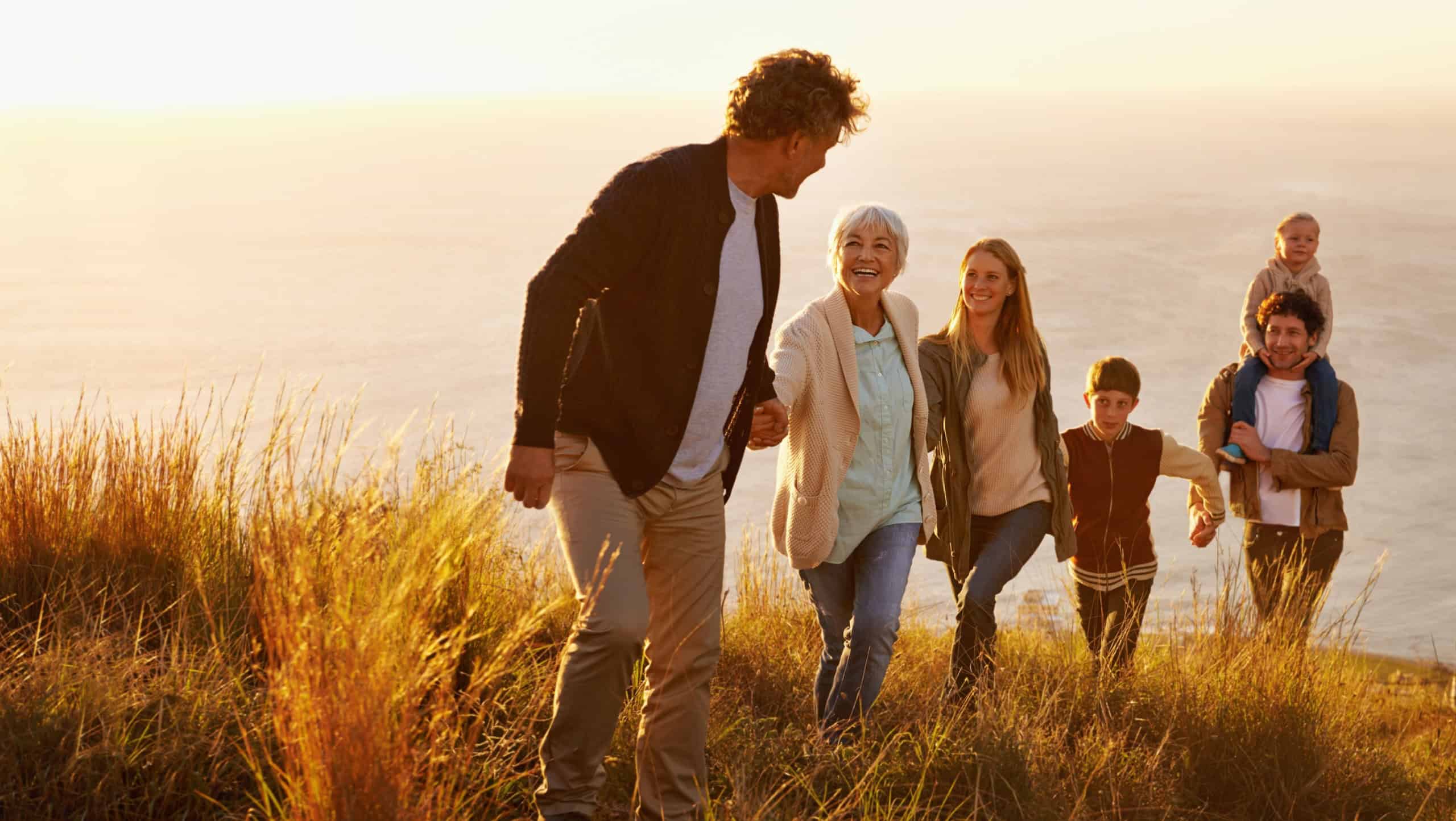 Follow the leader. A multi-generational family walking up a grassy hill together at sunset with the ocean in the background.