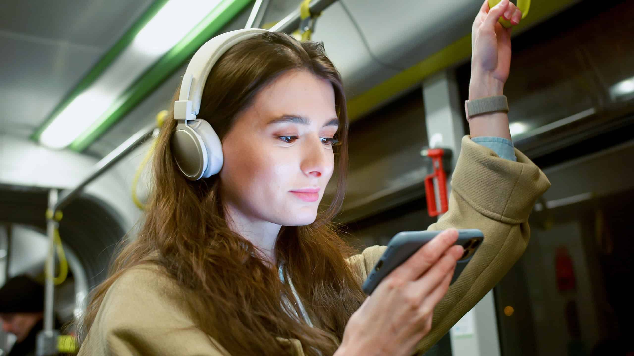 Side-view portrait of focused cute woman watching video on smartphone using headphones riding bus. Attractive happy caucasian female standing in public transport holding handrail.