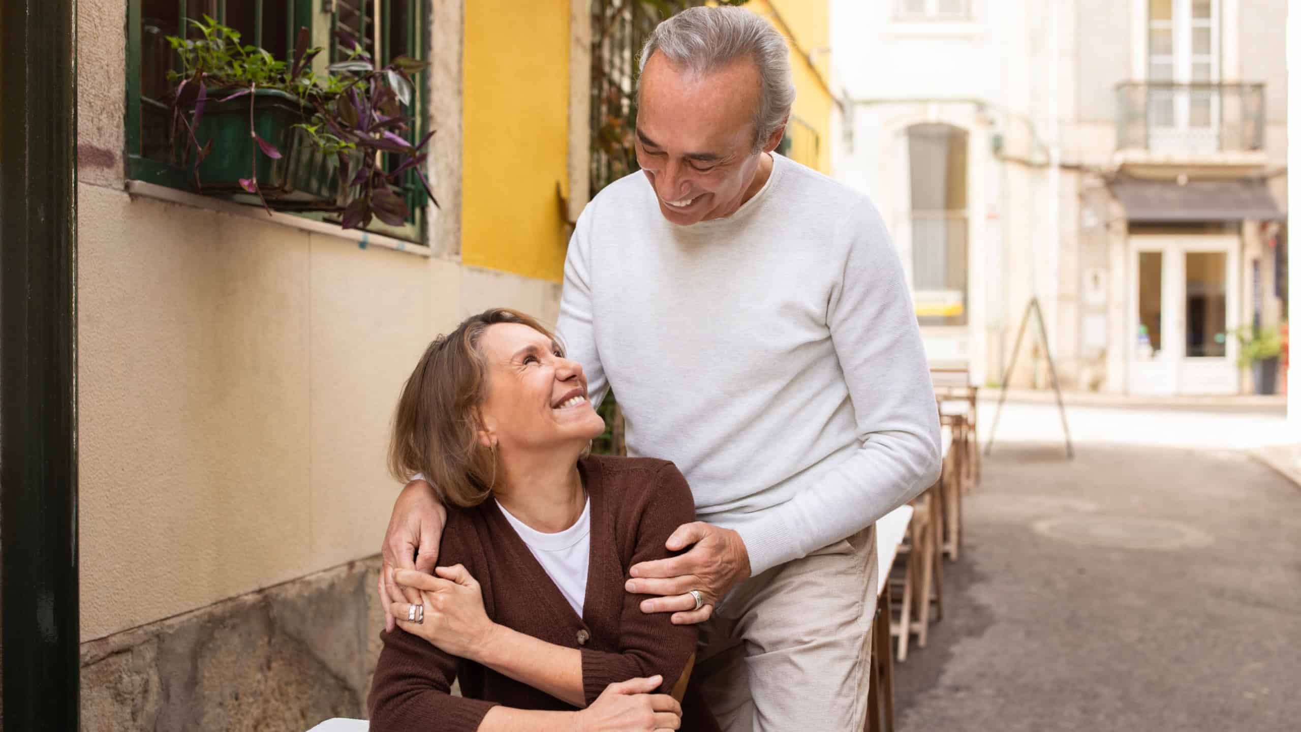 Cheerful Senior Spouses Sitting At Table Having Romantic Date In Outdoor Cafe During Vacation In Europe, Husband Hugging Wife Standing Behind Her Chair. Retirement Travel And Romance Concept