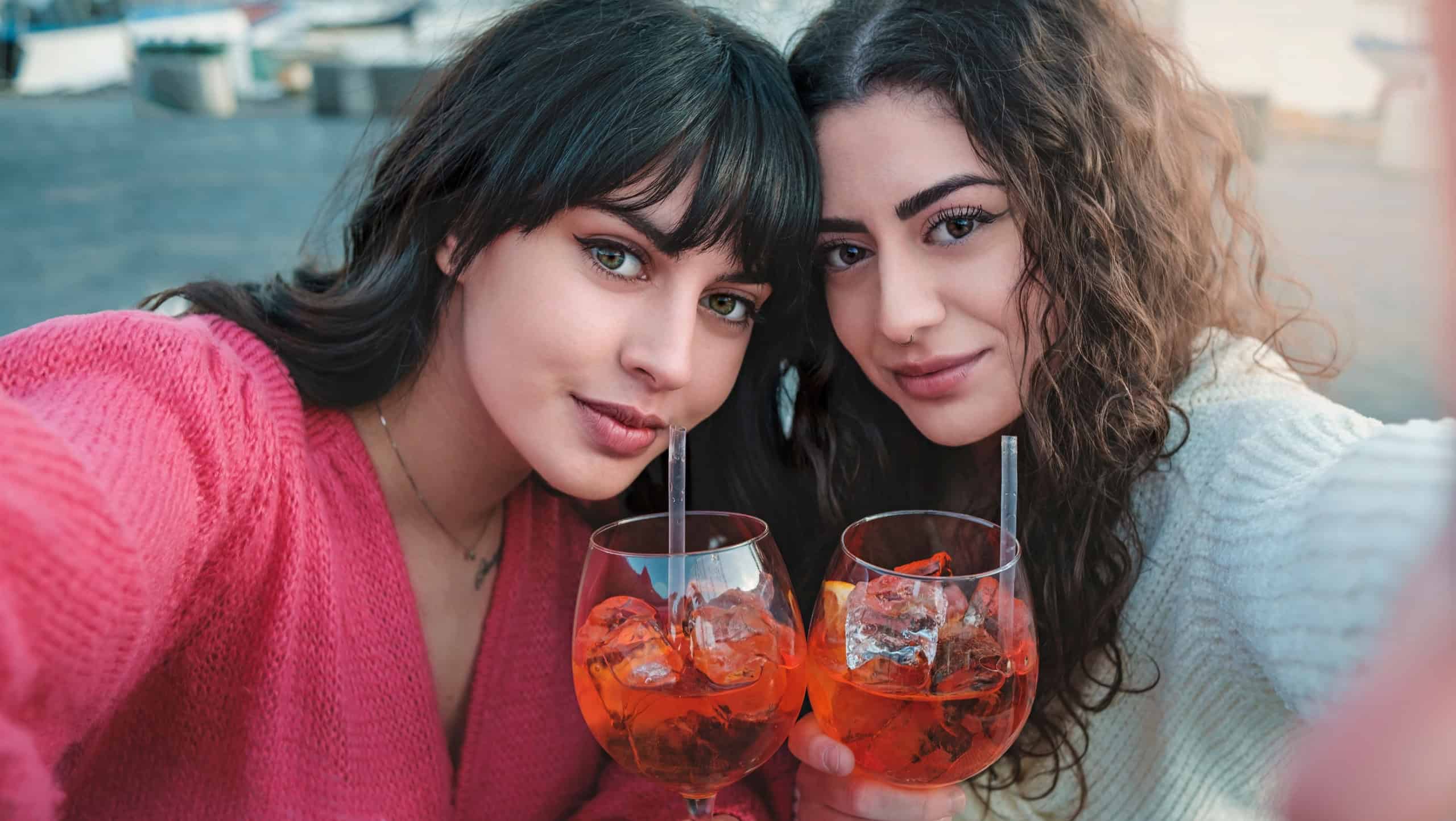 Two Caucasian girls (21 years old) taking a selfie with a spritz in hand, with a pier in the background. Creative shot simulating the women holding a smartphone for the photo.