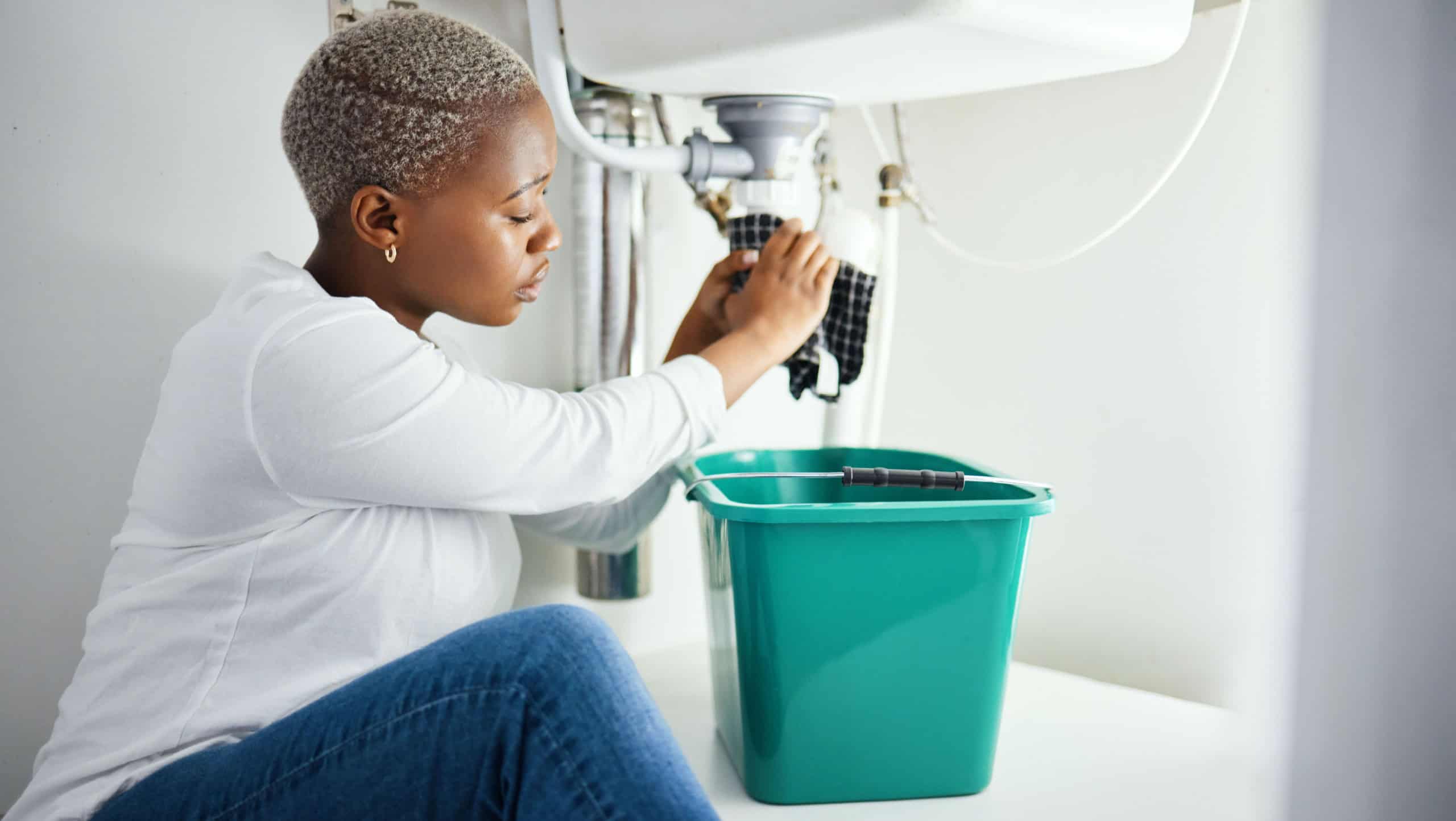 Plumbing, leak and a black woman in the bathroom of her home with a cloth and bucket waiting for assistance. Sink, emergency and a burst pipe with a young female homeowner in her house to stop water
