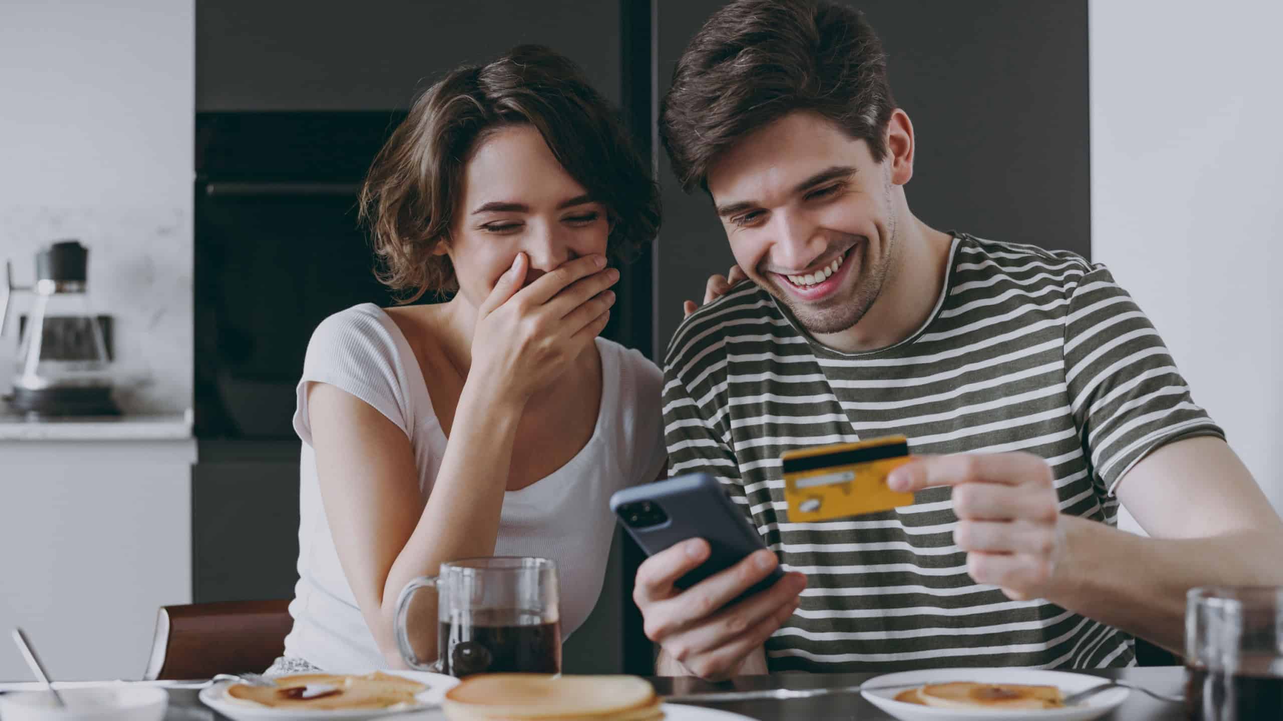 Young happy couple two woman man in casual clothes sit by table eat pancake use mobile cell phone credit bank card shop online order delivery in light kitchen at home together Healthy diet concept.
