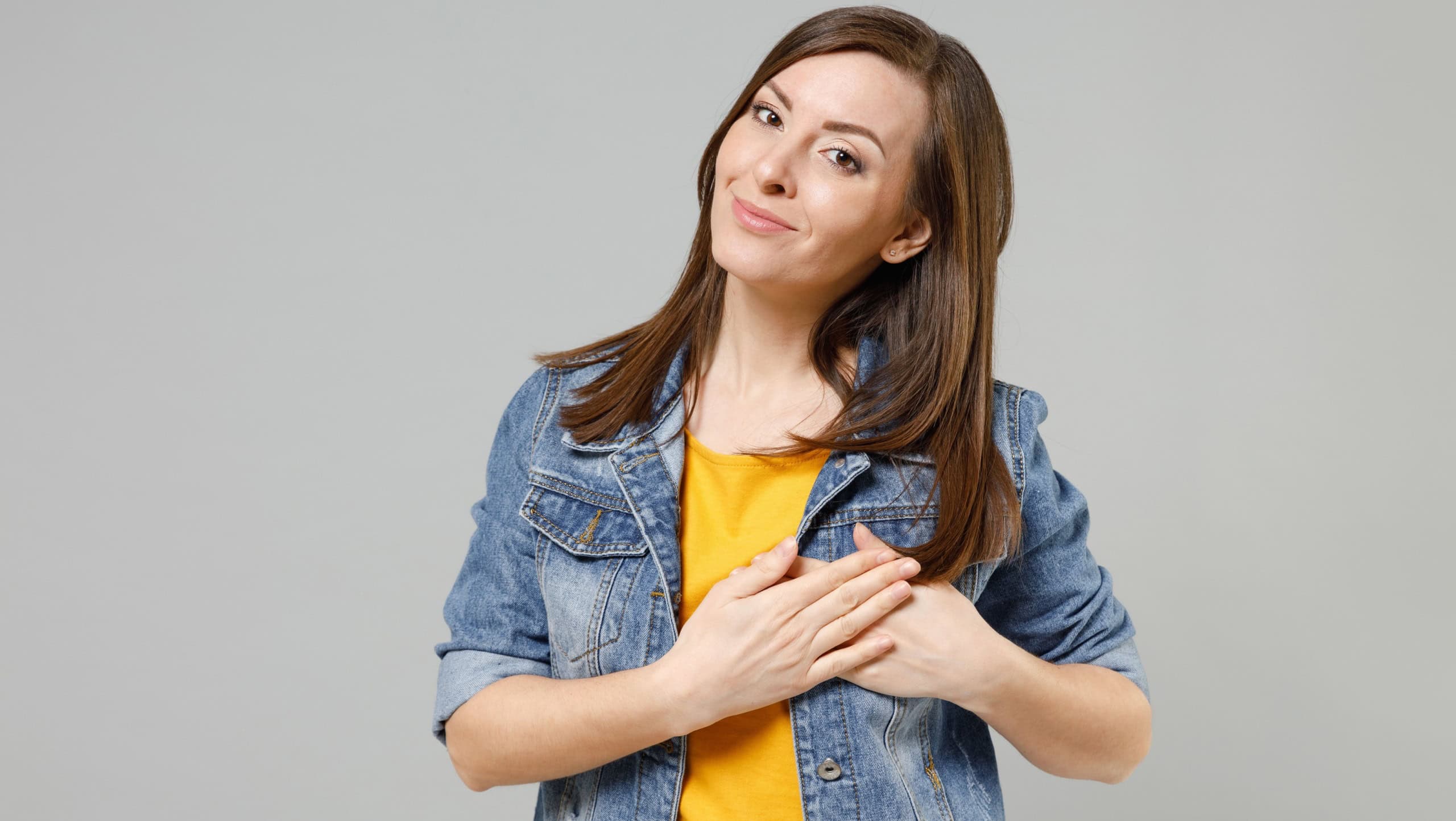 Young kind-hearted smiling happy caucasian woman 20s in casual trendy denim jacket yellow t-shirt put folded hands on heart isolated on grey color background studio portrait. People lifestyle concept.