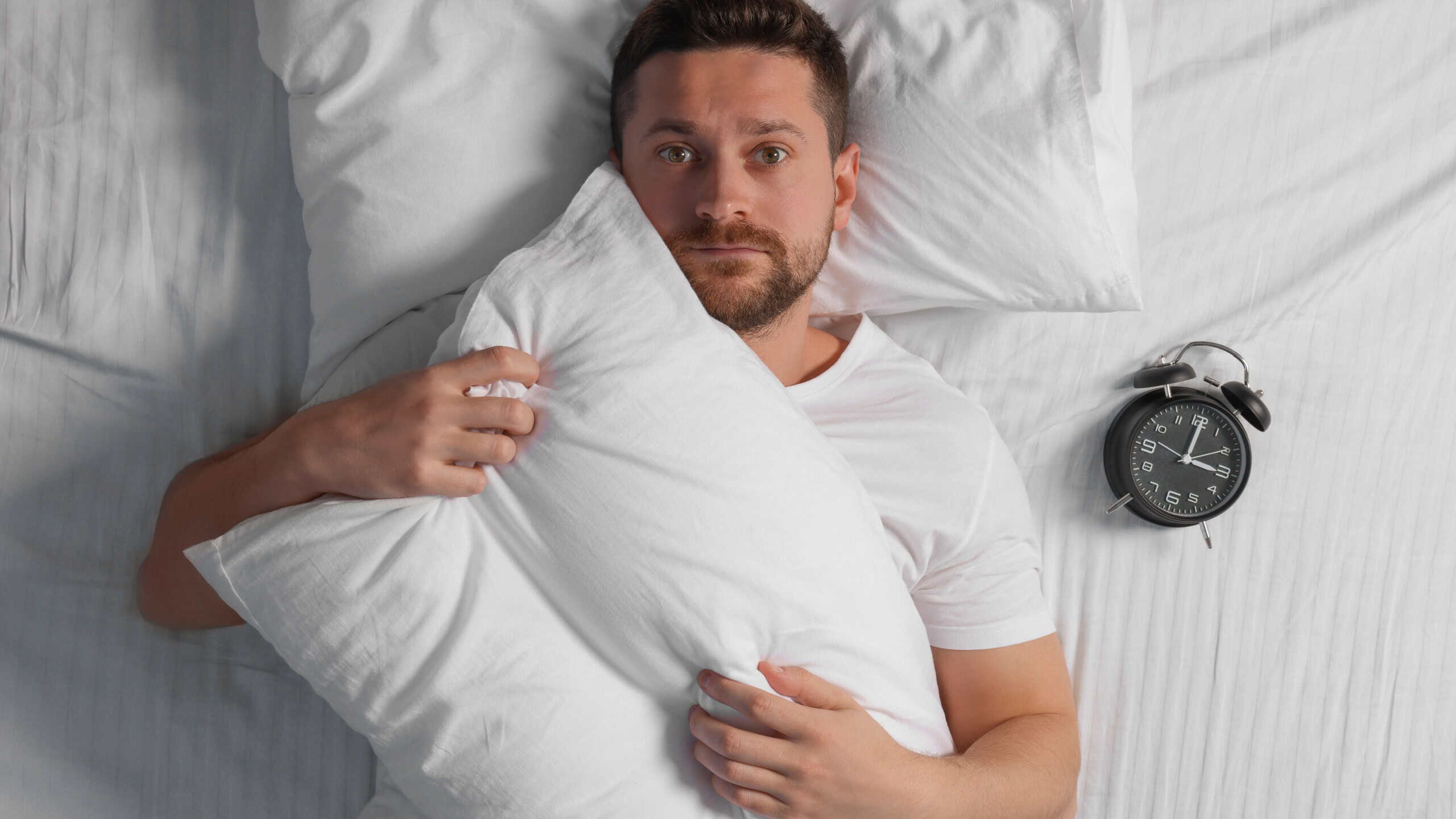 Sleepy man with pillow and alarm clock in bed, top view. Insomnia problem