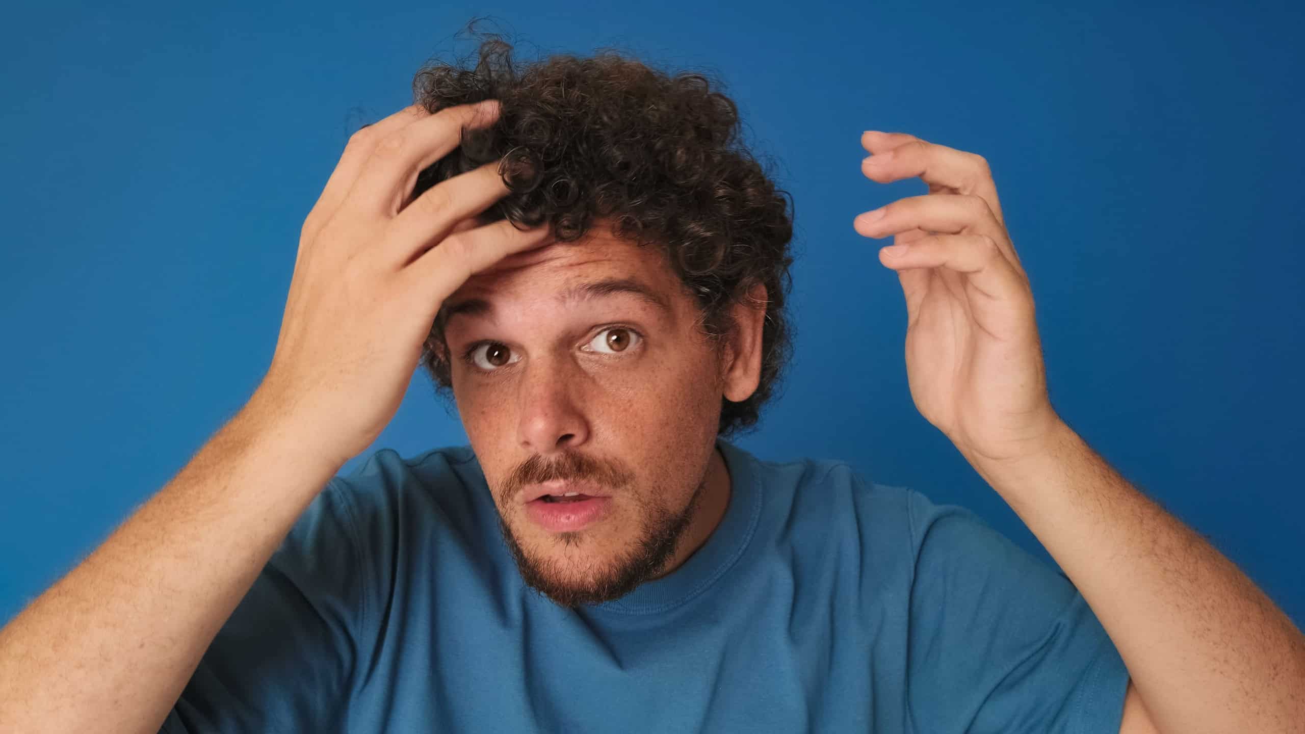 Close-up of guy with curly hair dressed in blue t-shirt, looking at the camera as if in mirror, fixing his hair on blue background in the studio