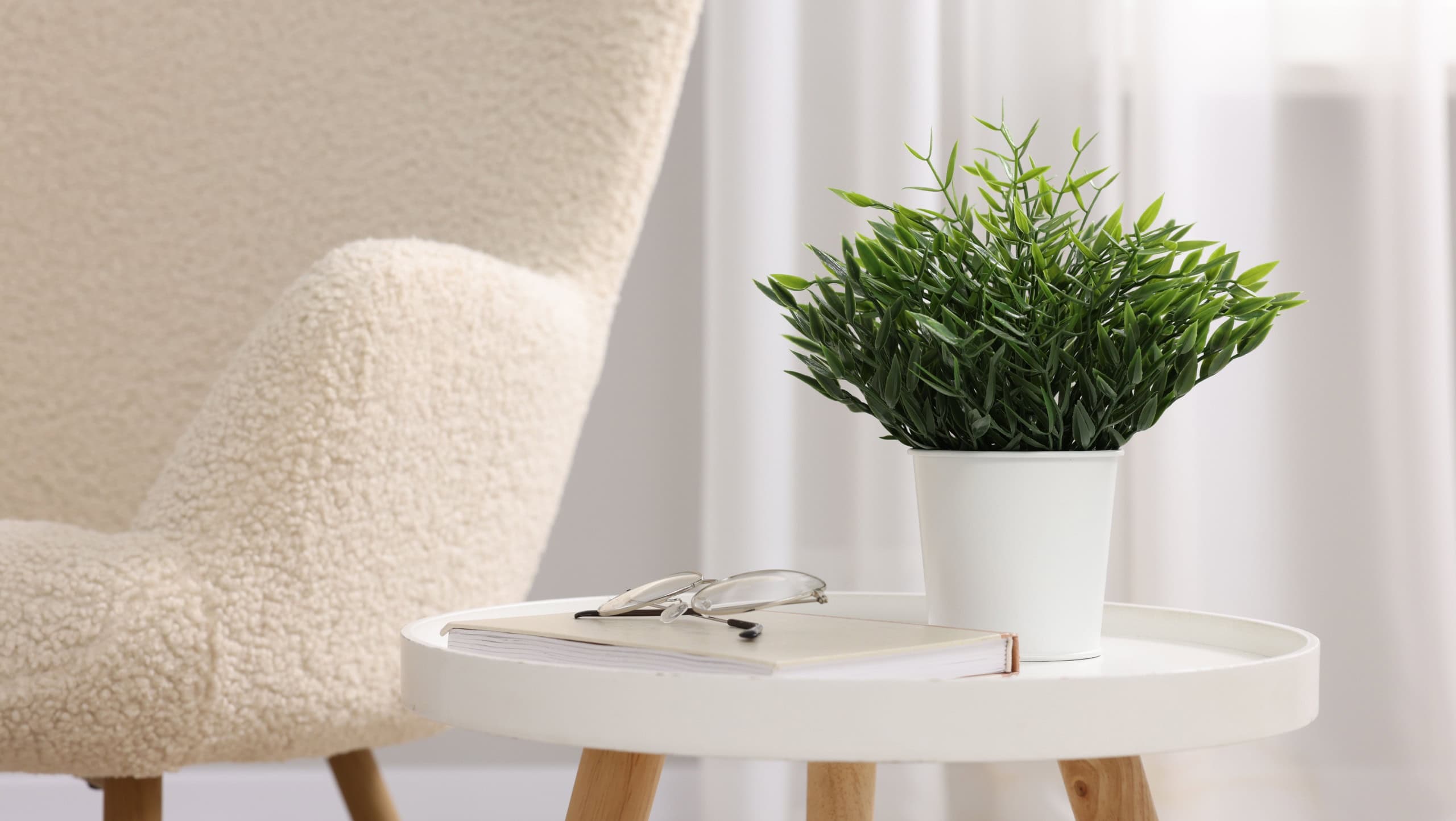Potted artificial plant, book and glasses on side table near armchair indoors