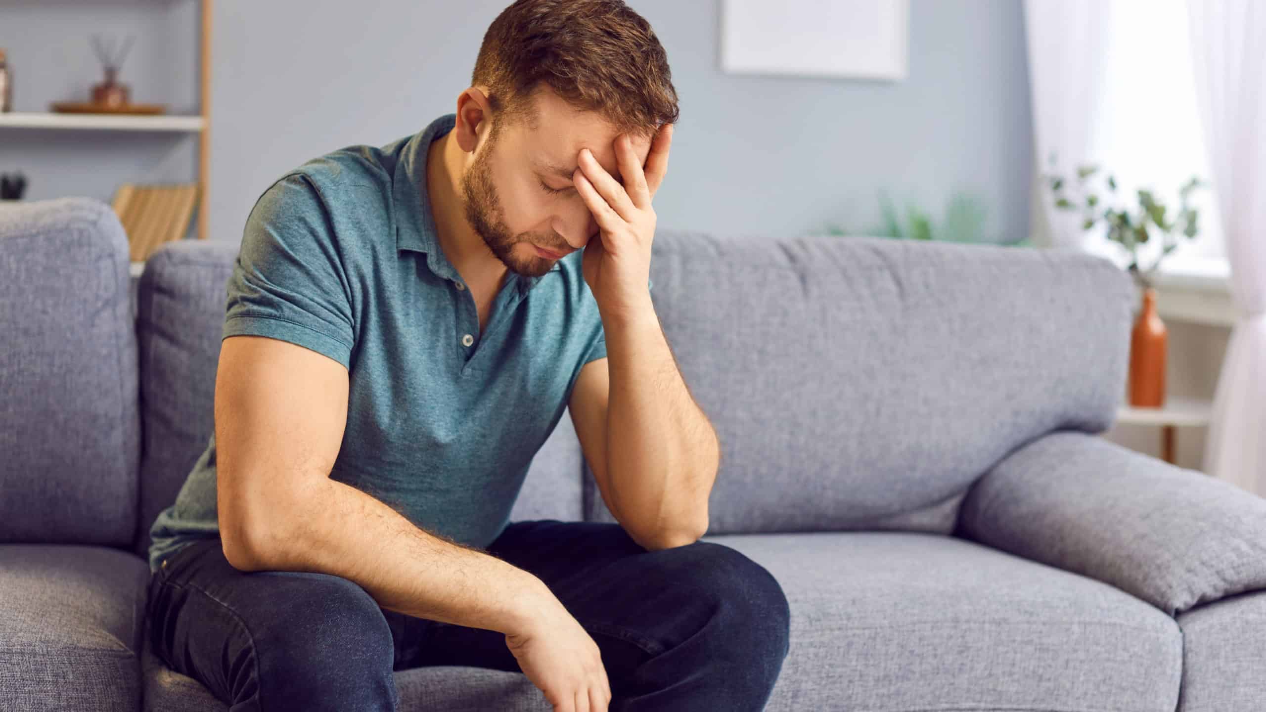 Portrait of upset sad depressed man sitting on sofa in living room at home alone suffering from headache. Young guy in bad thoughts feeling lonely indoors or having problems in relationships