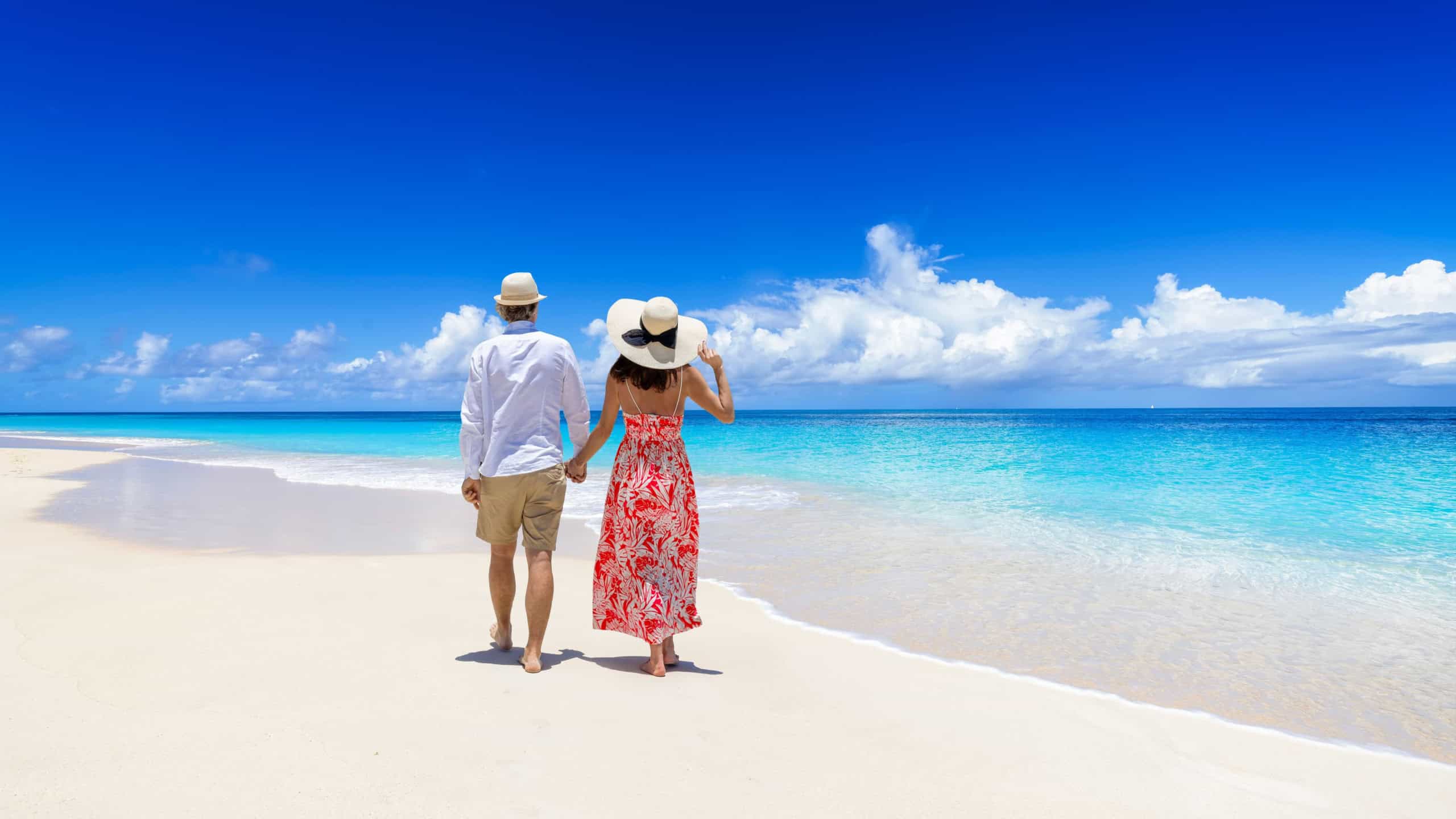 Happy holiday couple with sunhats walks down a tropical paradise beach with turquoise sea and golden sand
