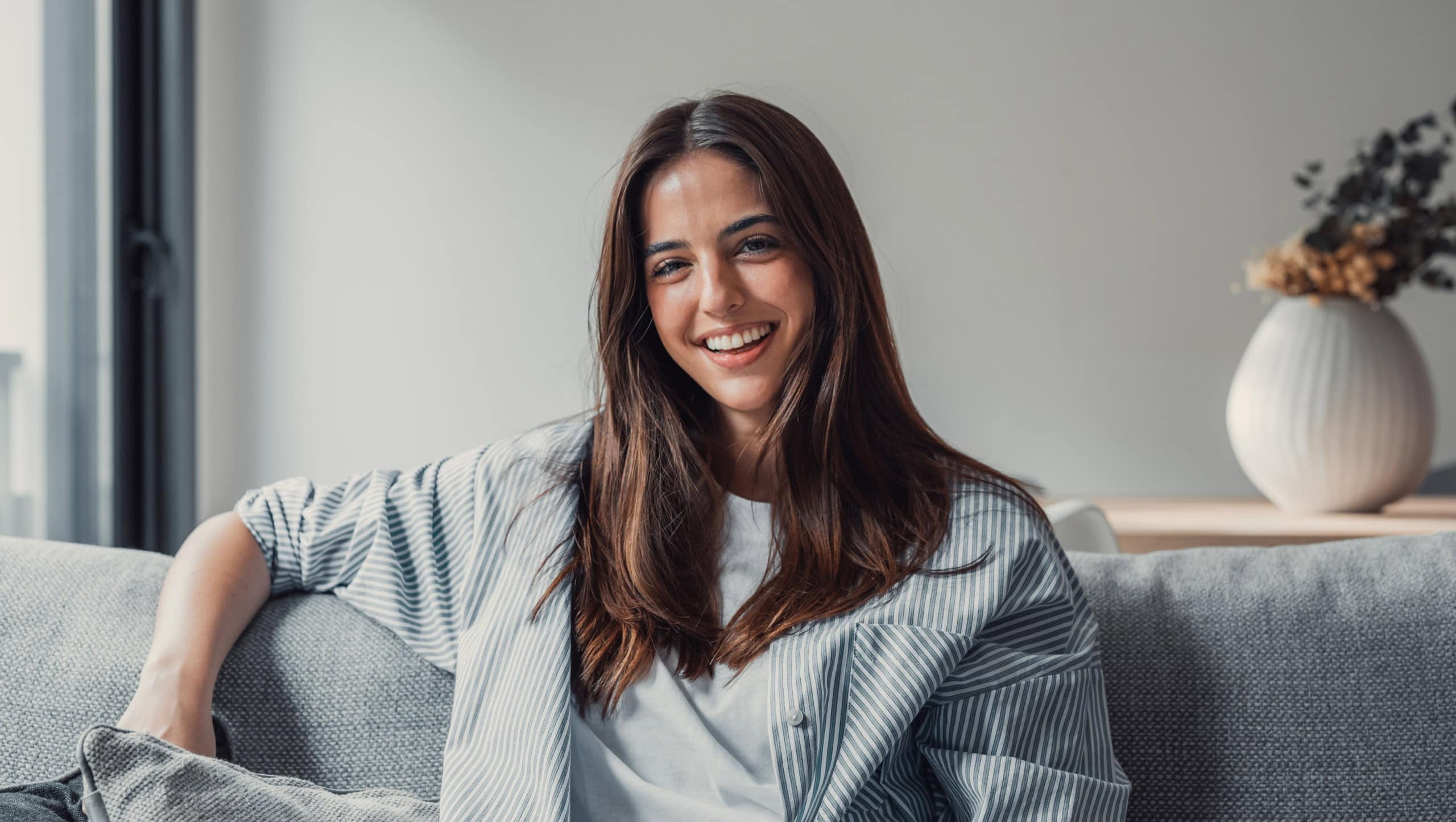 Happy attractive young adult woman girl sitting on sofa looking at camera in modern cozy home, smiling single 20s lady beautiful face relaxing on couch alone enjoy wellbeing posing for portrait