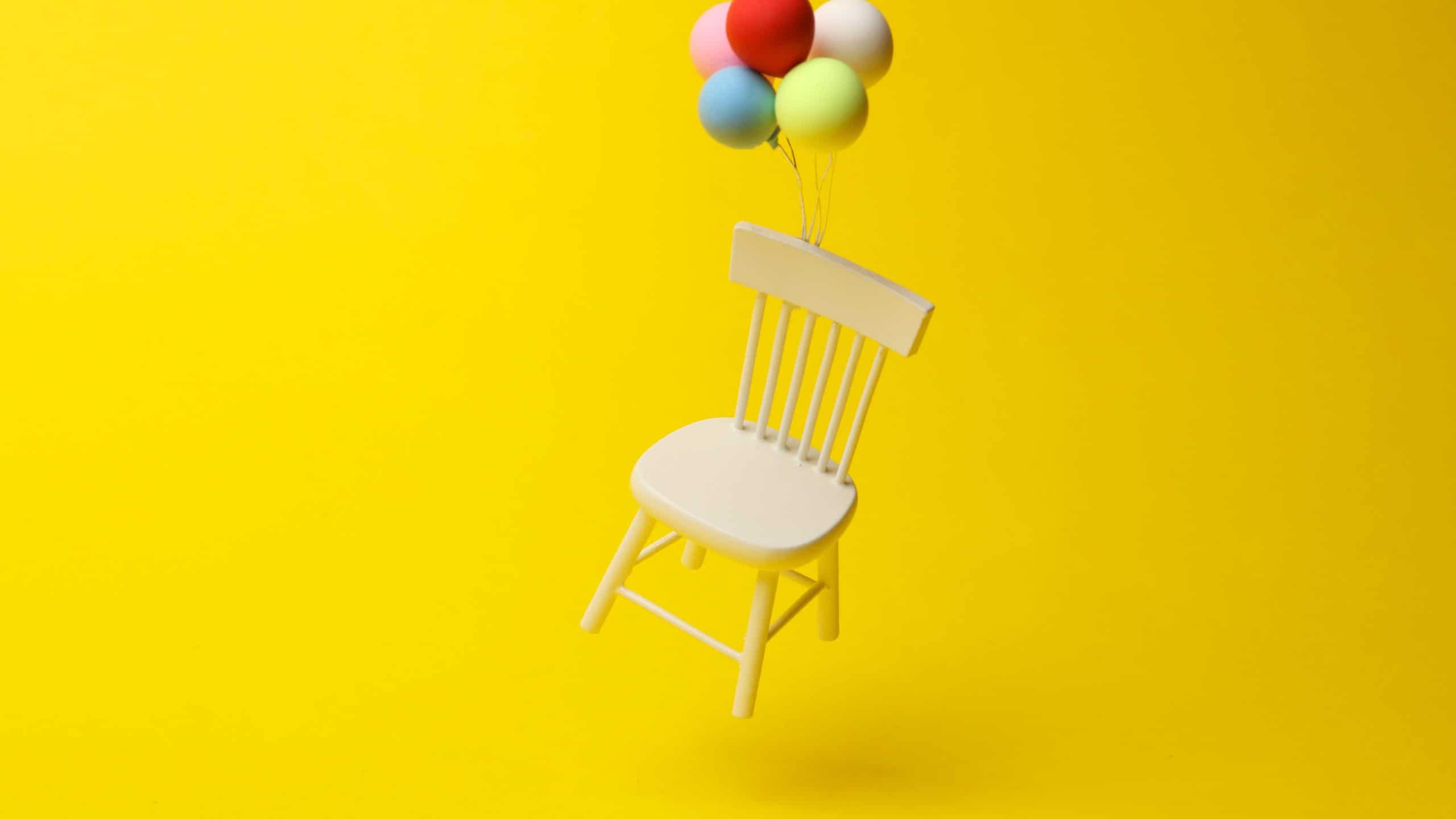 White miniature chair tied to balloons floating on a yellow background. Minimalism.