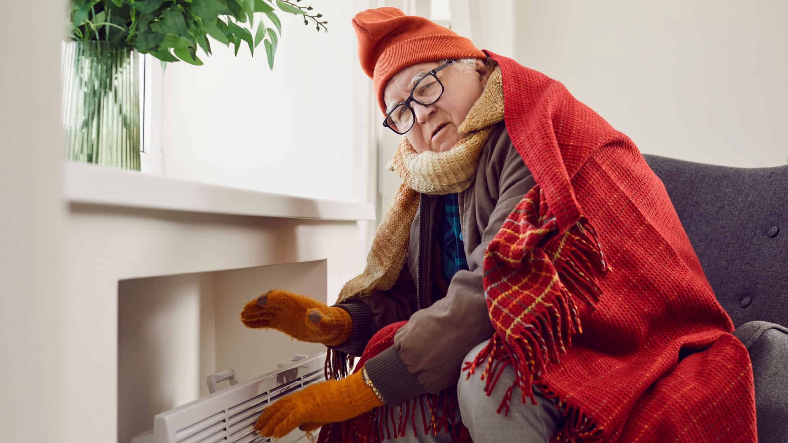 Portrait of a worried sad senior elderly man looking at camera wrapped in a blanket and trying warming up on electric heater. Mature man freezing at home. Heating problems concept.