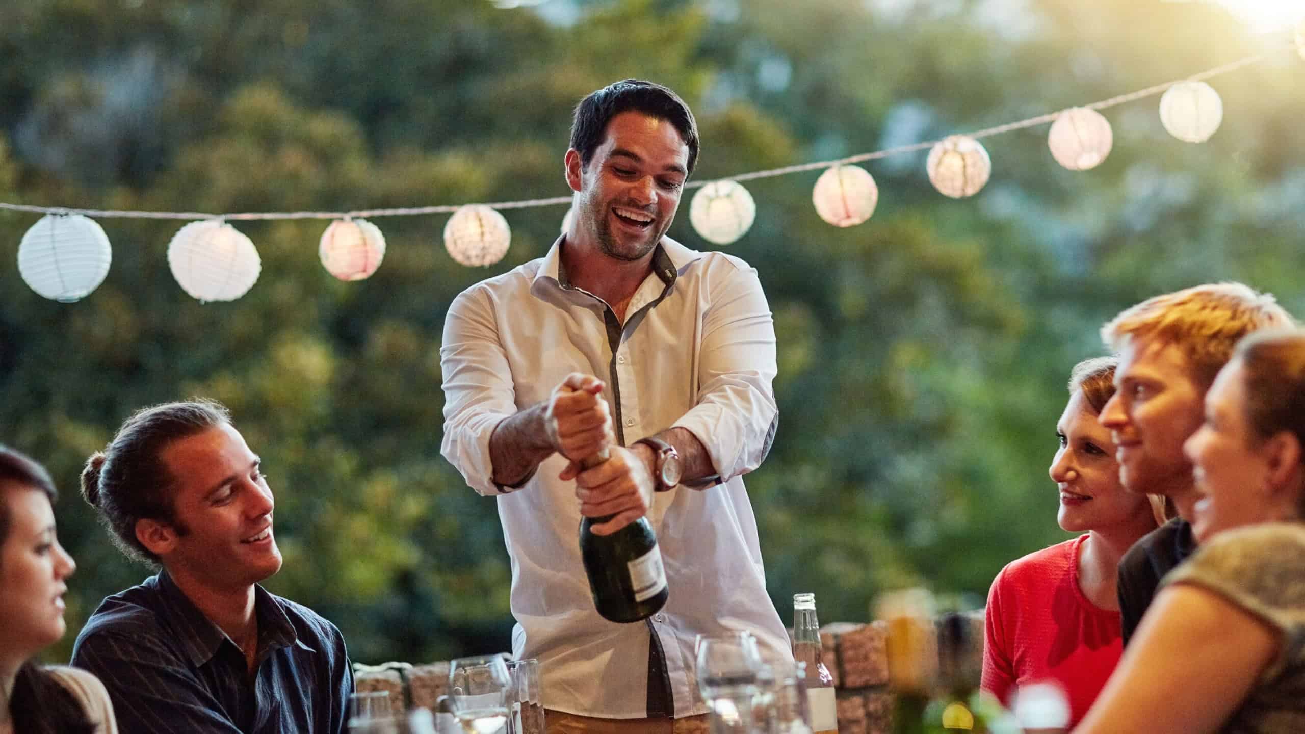 Champagne, open bottle and friends at party for Octoberfest, dinner and celebration in Munich. Wine, group and happy people at reunion for social gathering or excited host serve alcohol drink outdoor