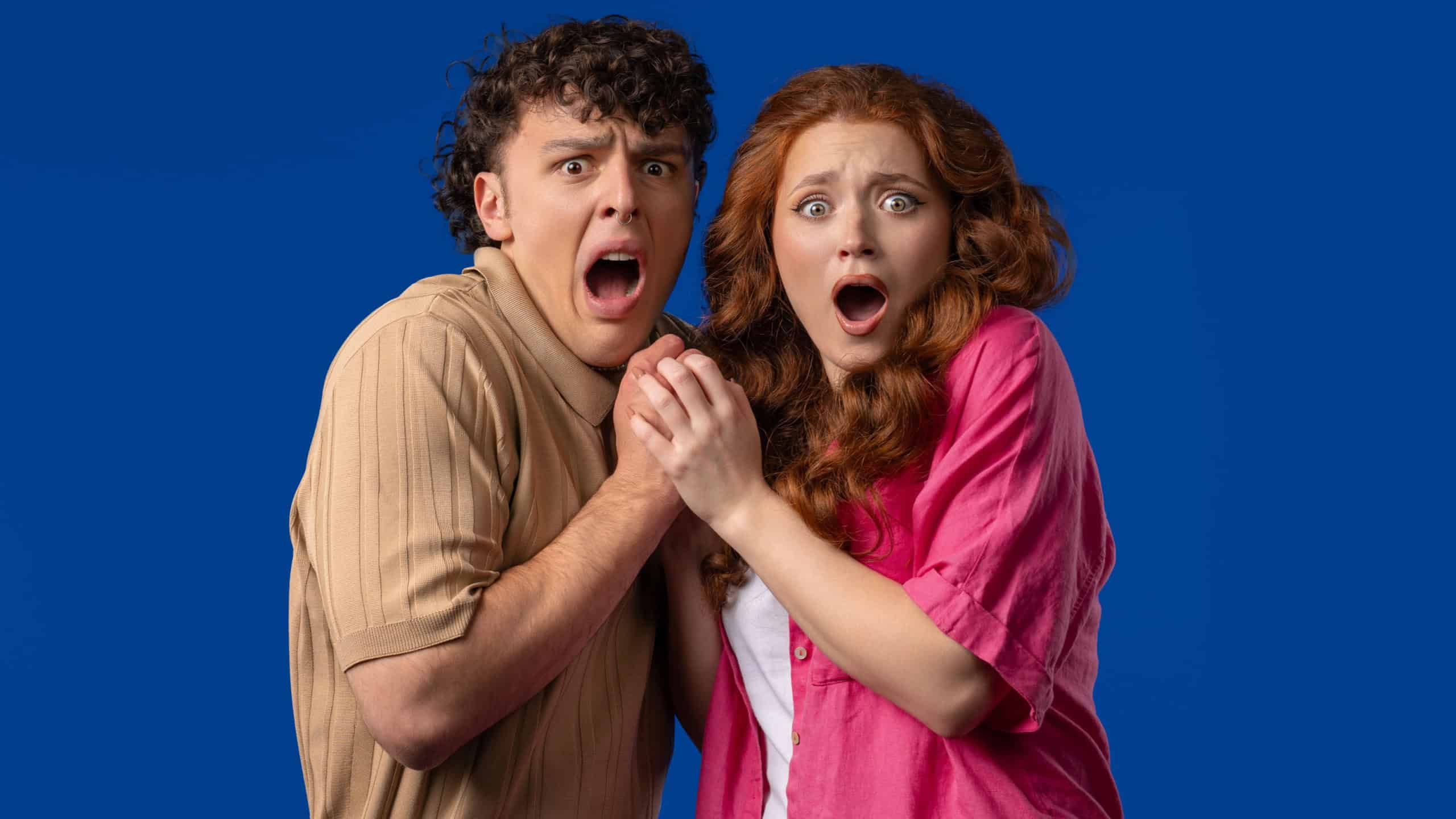 Frightened white american couple afraid of something and looks into camera with big eyes full of horror on blue wall background. Red haired woman and man. High quality