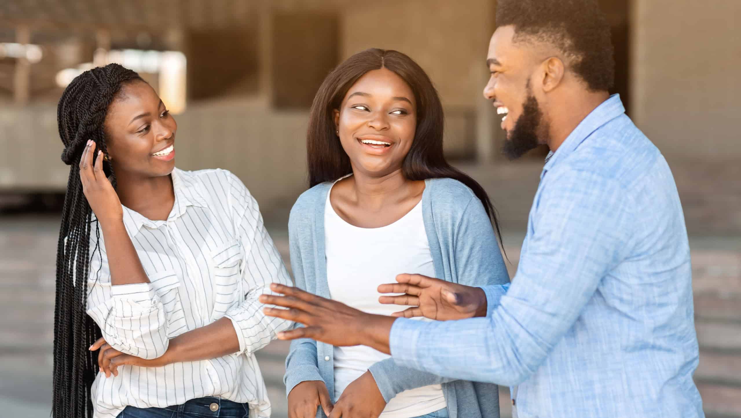 Three happy afro friends chatting and laughing outdoors in the street, handsome guy telling joke to girls, explaining something funny