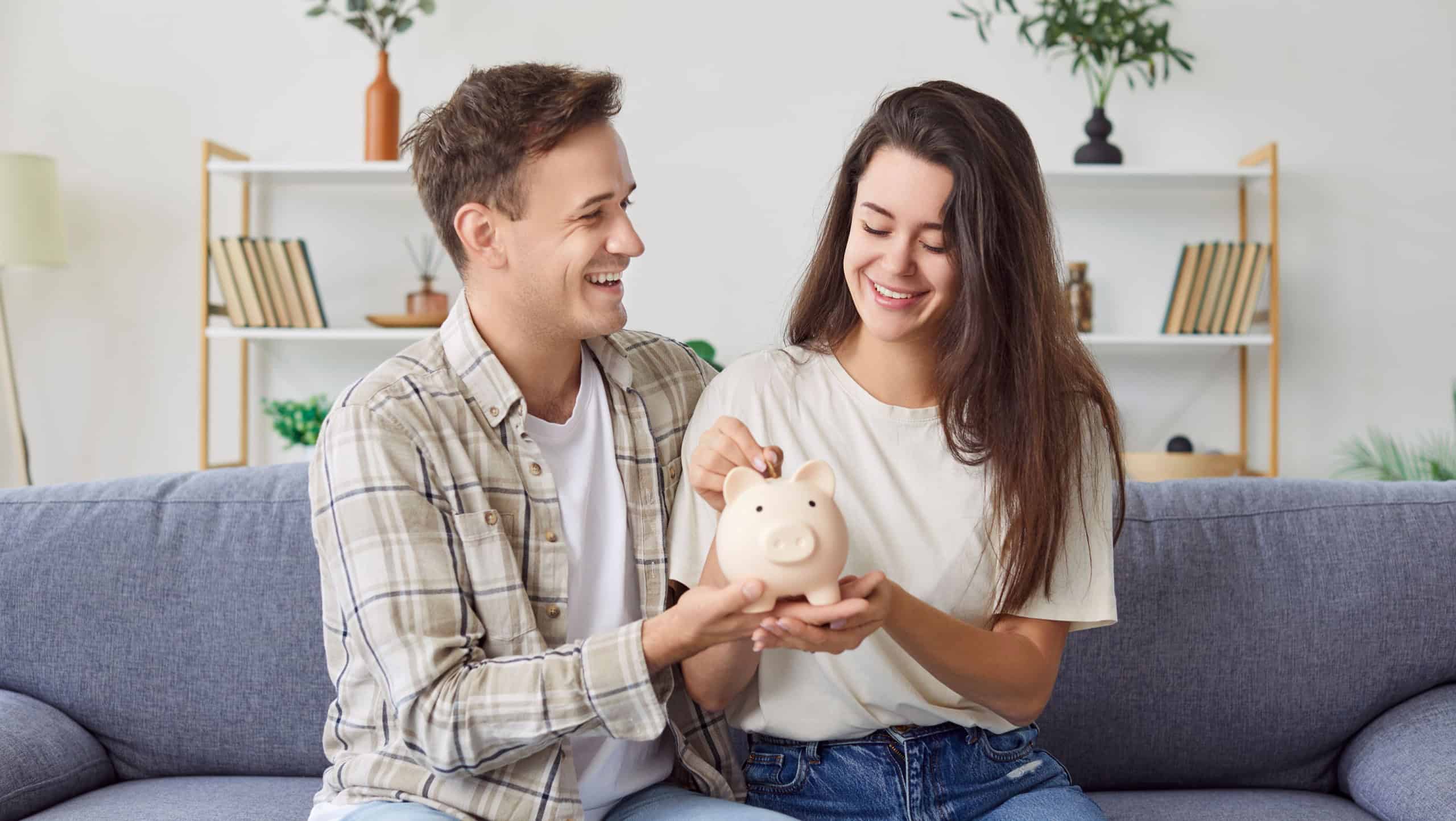 Young family couple sitting on couch, holding piggy bank in their hands, planning their budget together. Happy woman putting coin in pink moneybox. Saving up, making plans, family budget management.