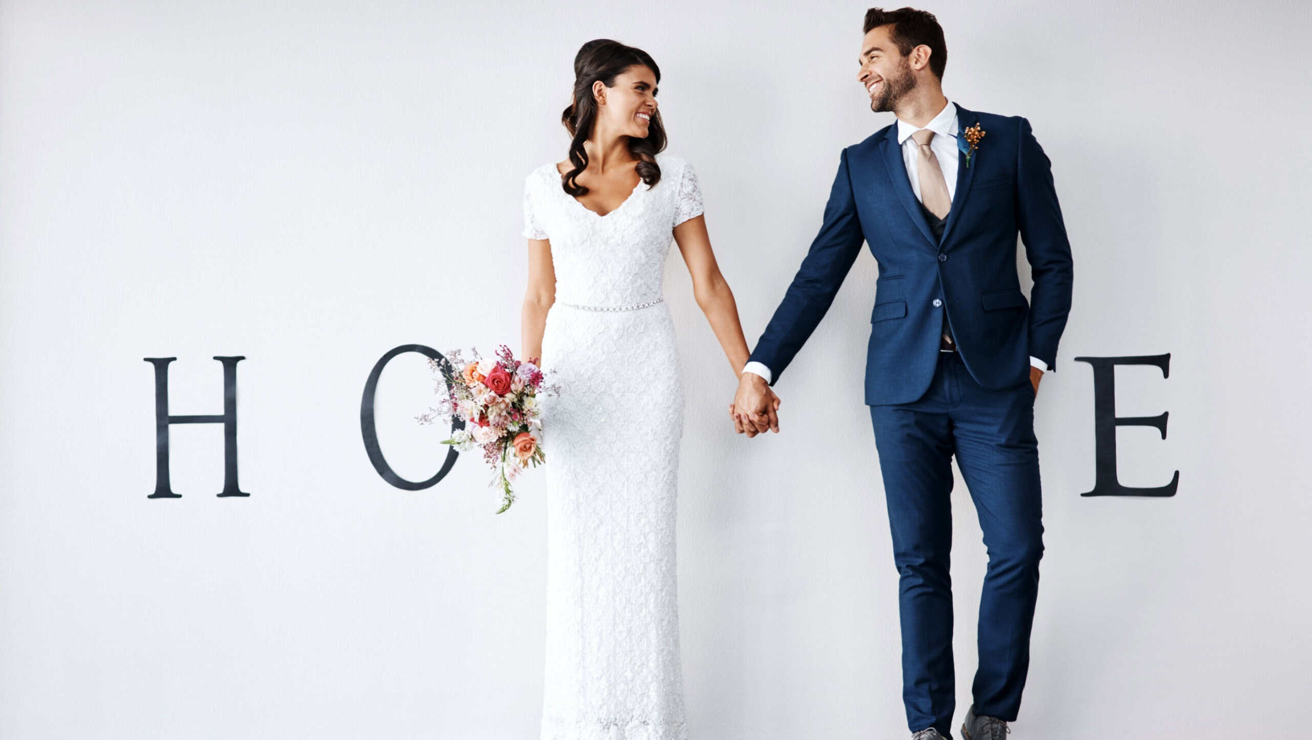 Smile, bride and groom holding hands in new home for support, love or bouquet. Newlywed couple, man or woman in house together for marriage, wedding or mortgage investment on white wall background