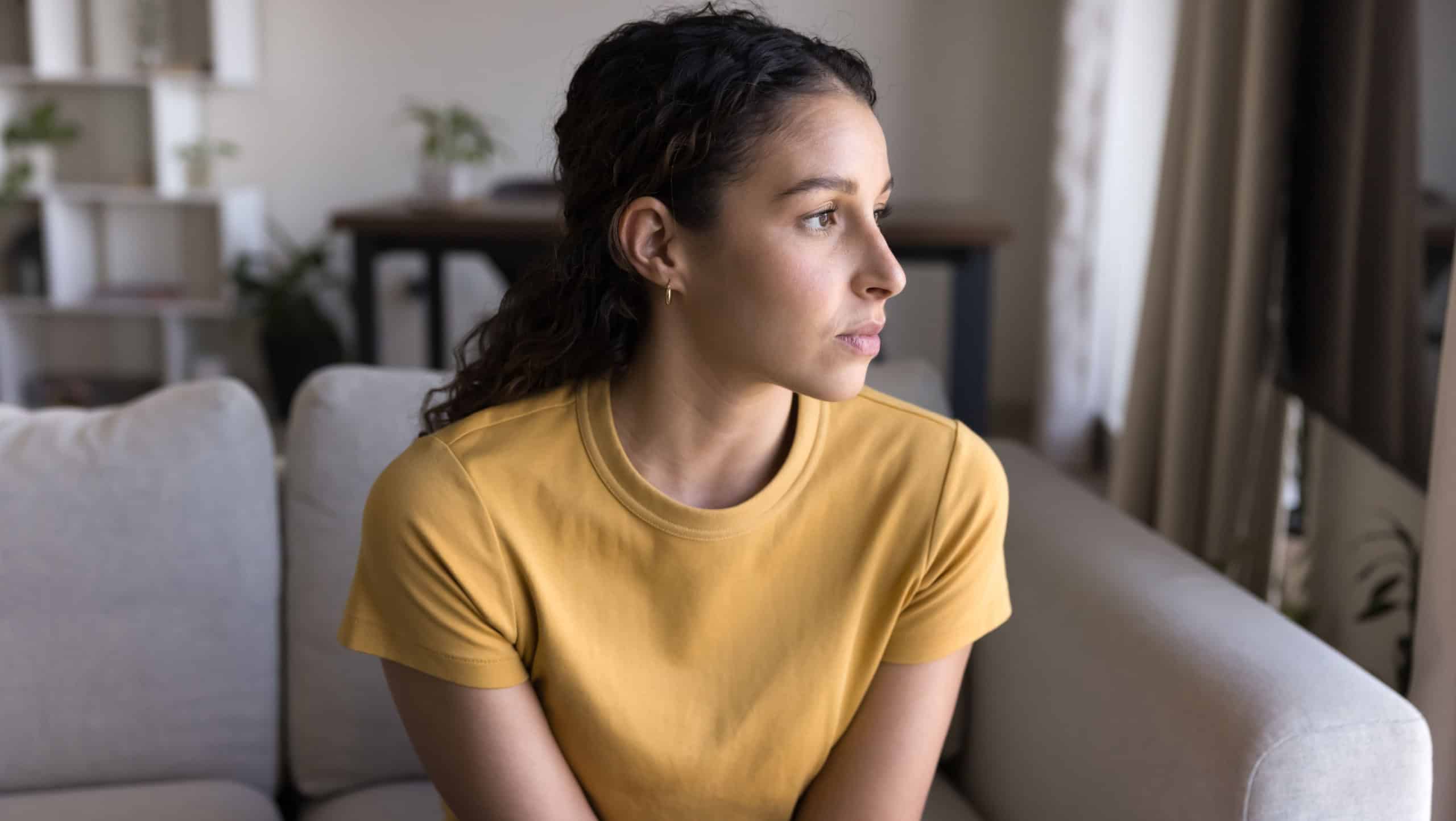 Serious thoughtful attractive 20s Latin woman looking at window away in deep thoughts, sitting on sofa, thinking on problem solving at home. Concerned pensive Hispanic young adult girl casual portrait