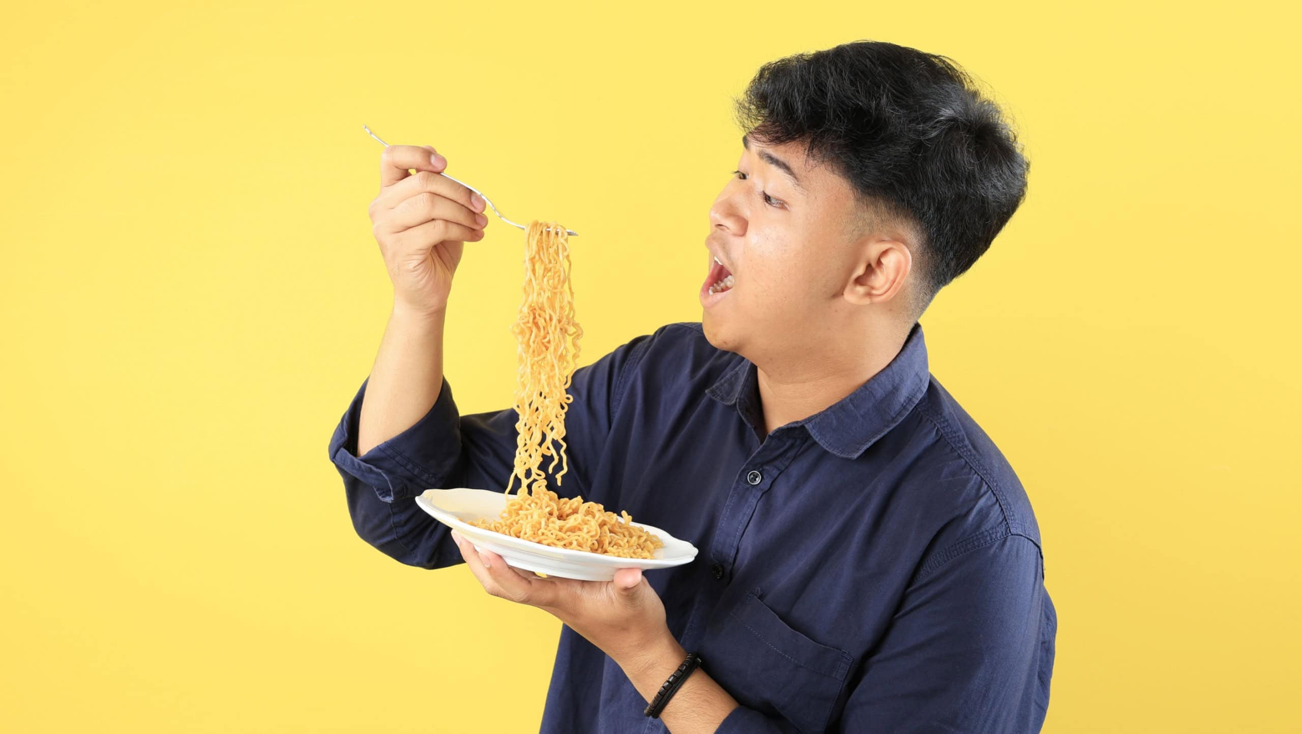 Young Asian Boy Student Eat Instant Noodle Fried Mie Goreng on White Plate, Isolated on Yellow Background