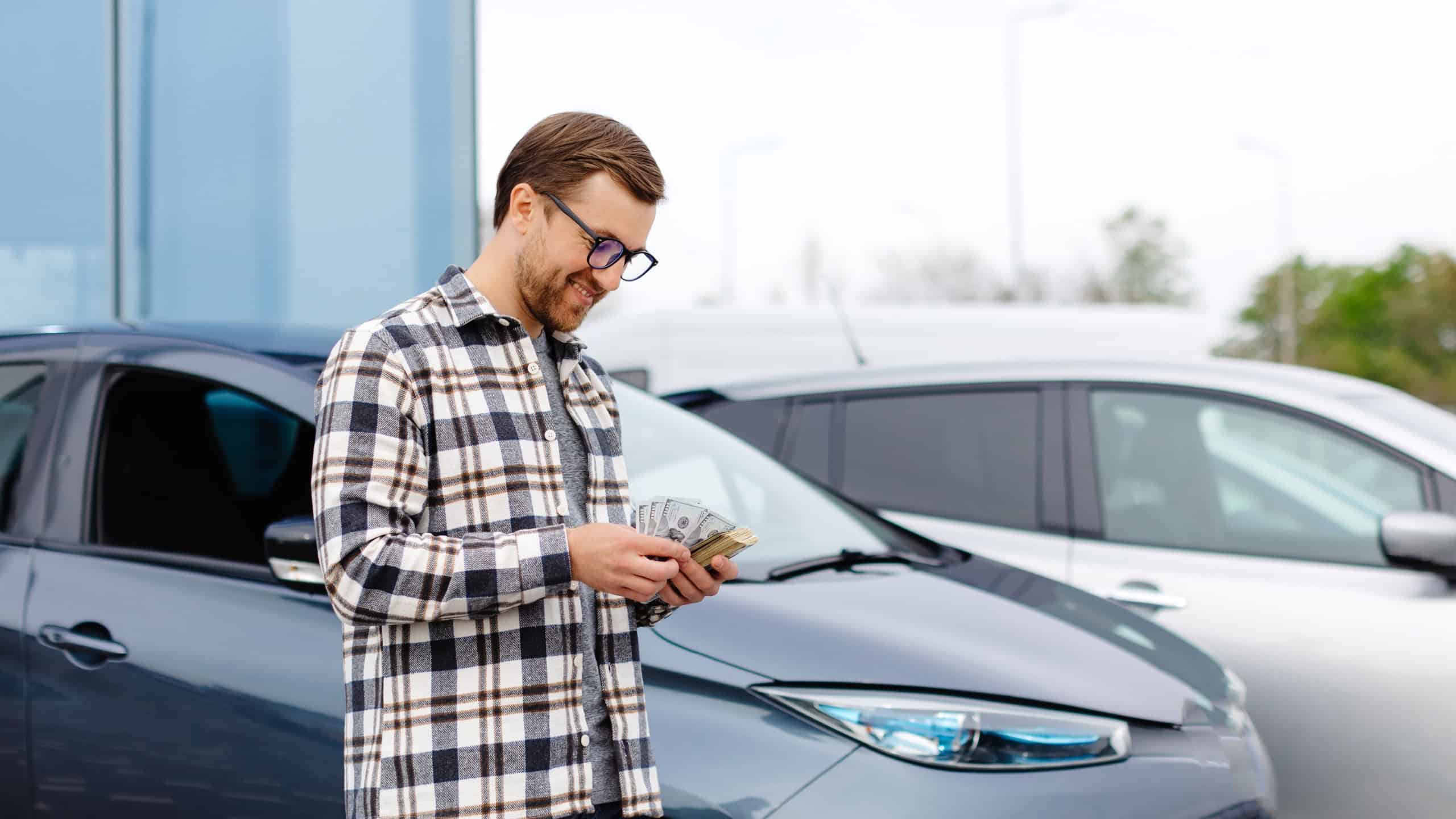 Young man holding money in hand standing front car and prepare pay by installments - insurance, loan and buying car concept insurance. Buy and down payment a car.